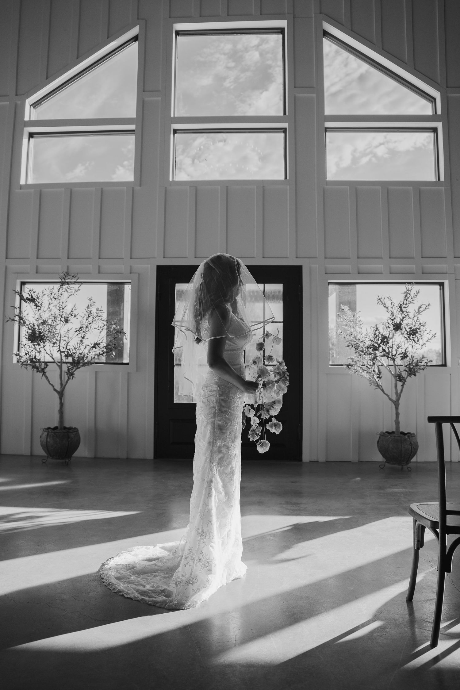 Black and white photo of bride in lace gown holding bouquet in sunlit barn venue.