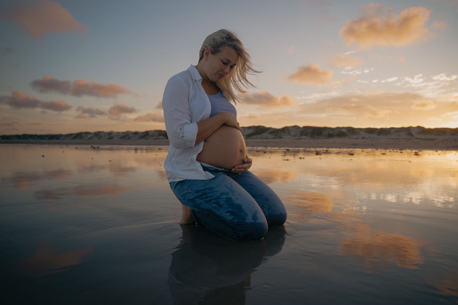 Pregnant woman kneels on reflective beach at sunset, cradling her baby bump in golden light.