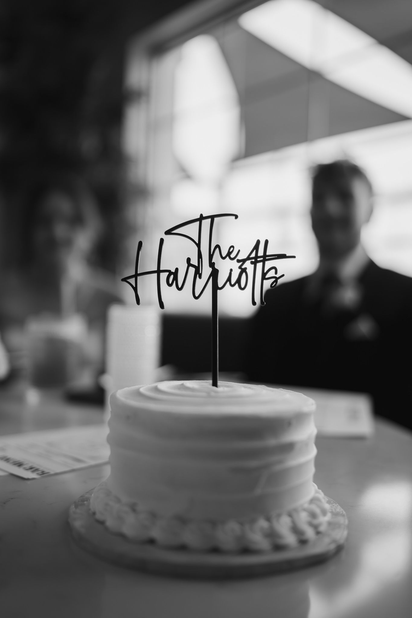 Black and white photo of wedding cake with The Harriotts cake topper at reception.