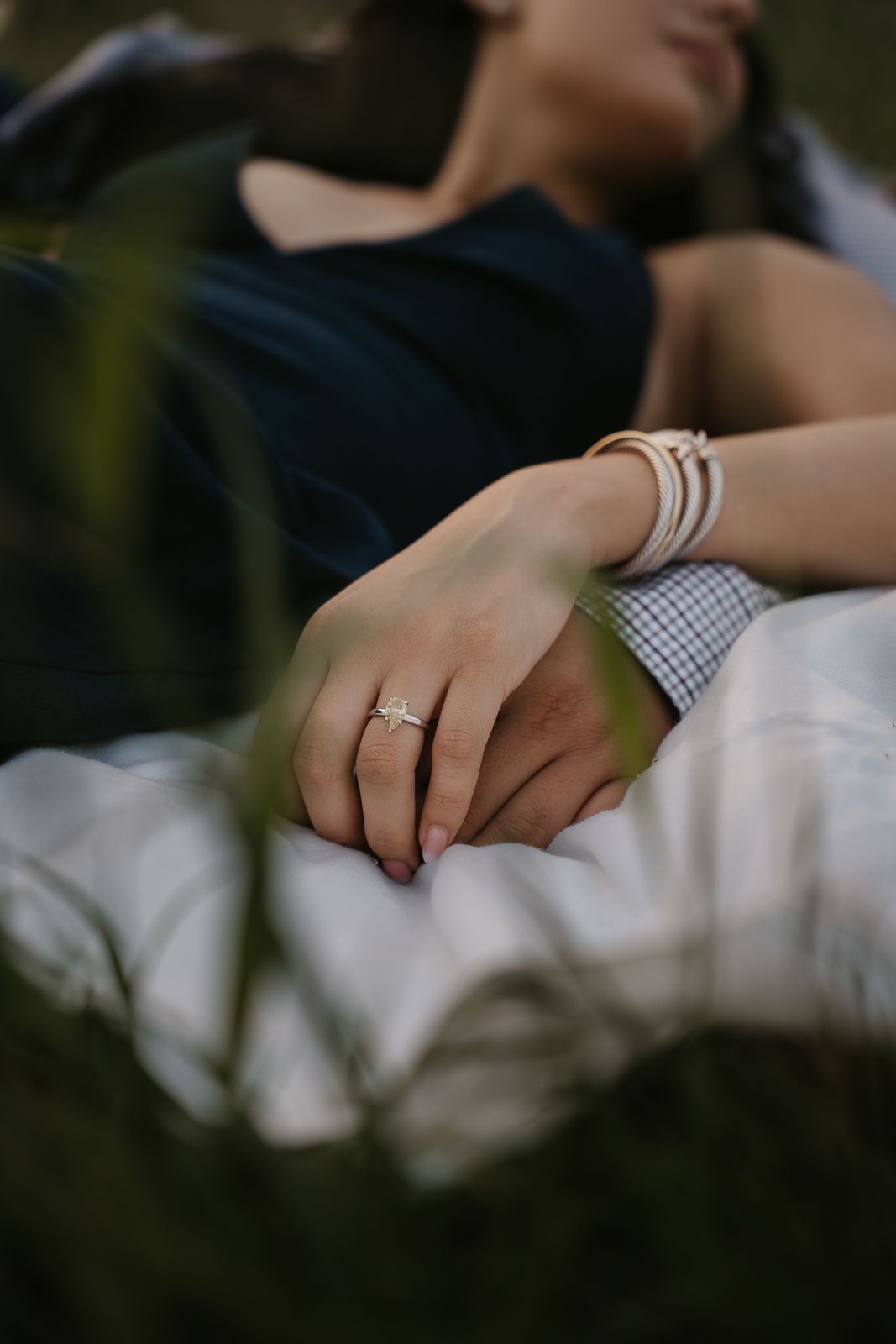 Close-up of a woman's hand displaying a diamond engagement ring and gold bracelets while resting outdoors.