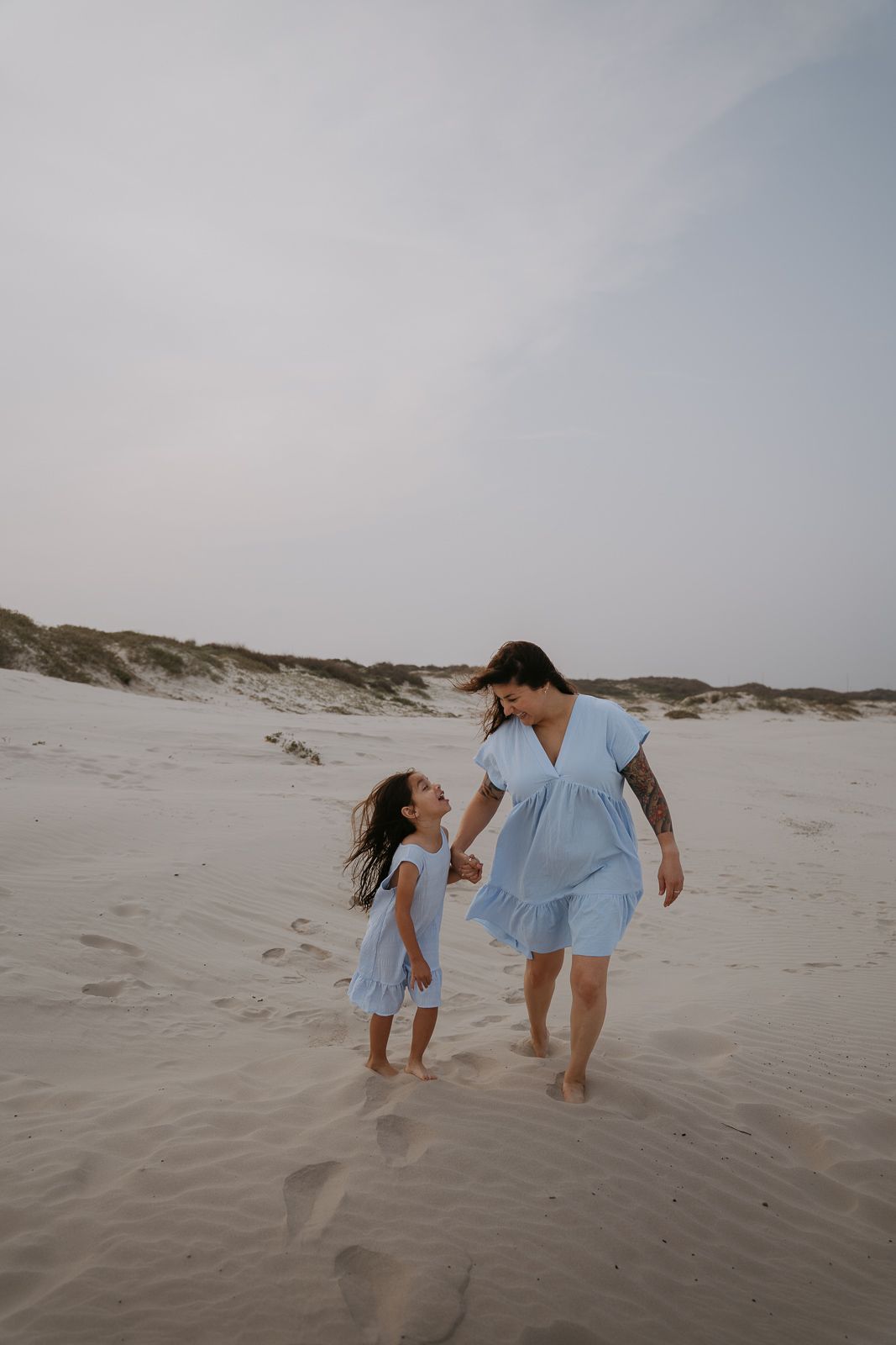 Mother and daughter in matching blue dresses walking on sandy beach dunes.