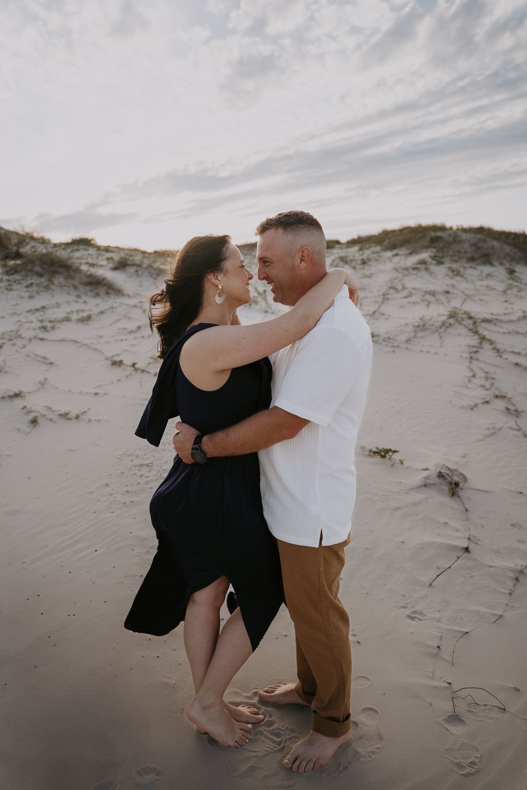 Happy couple embracing and smiling at each other barefoot on sandy beach at sunset.