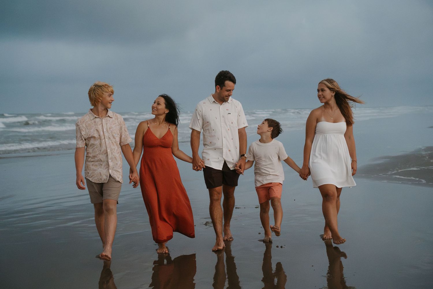 Family of five walking hand-in-hand on a beach with ocean waves, wearing casual summer outfits on a cloudy day.