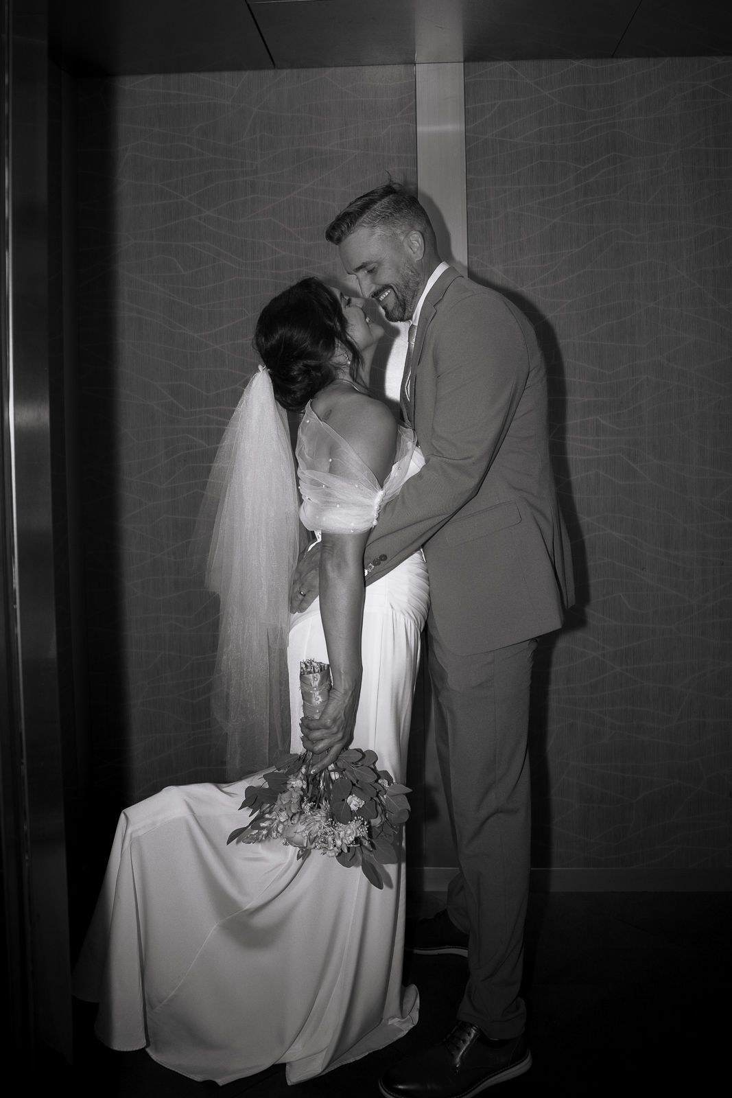 Black and white photo of bride and groom sharing a romantic kiss in an elevator on their wedding day.