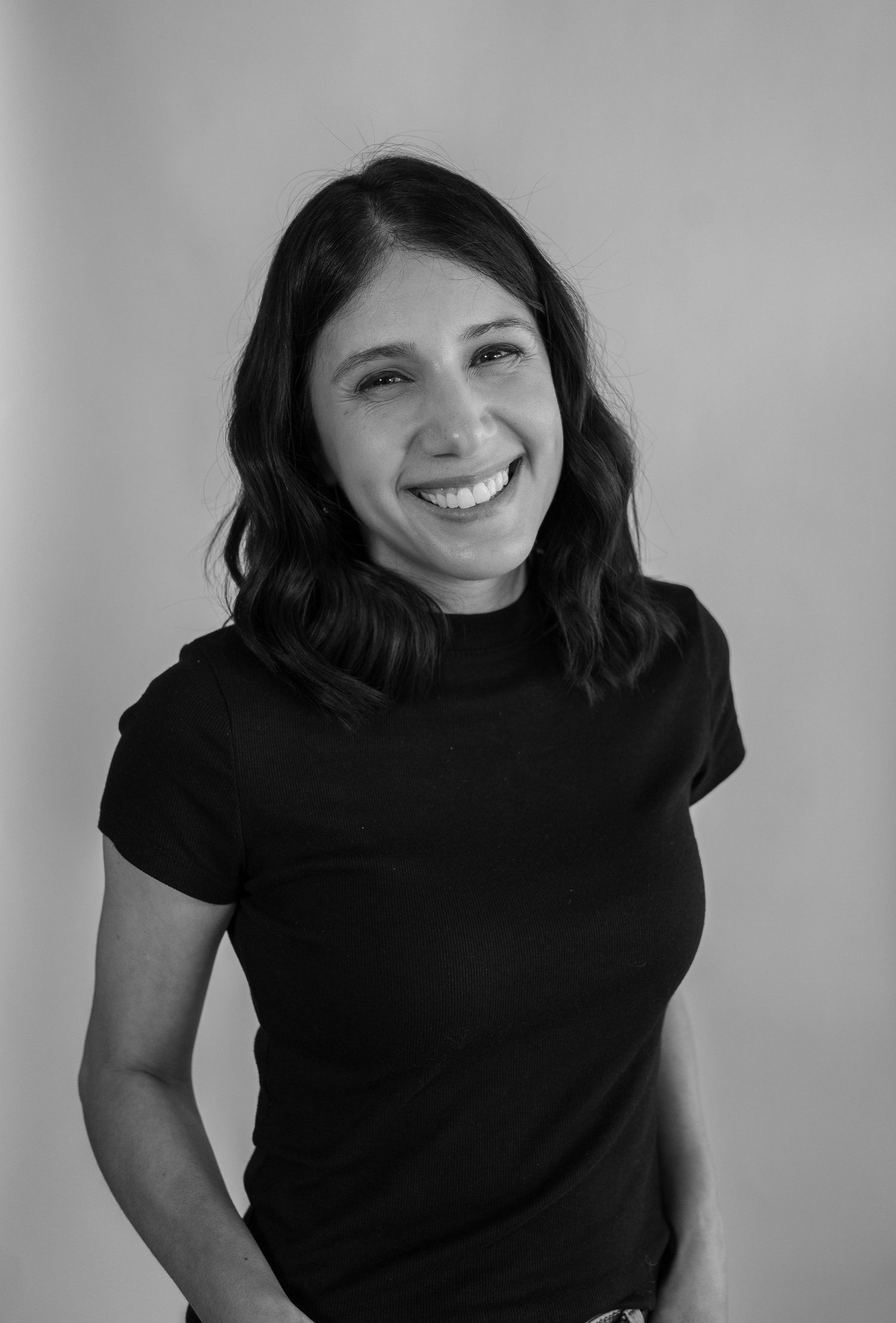 Smiling young woman with dark wavy hair wearing a black t-shirt in a black and white portrait photo.