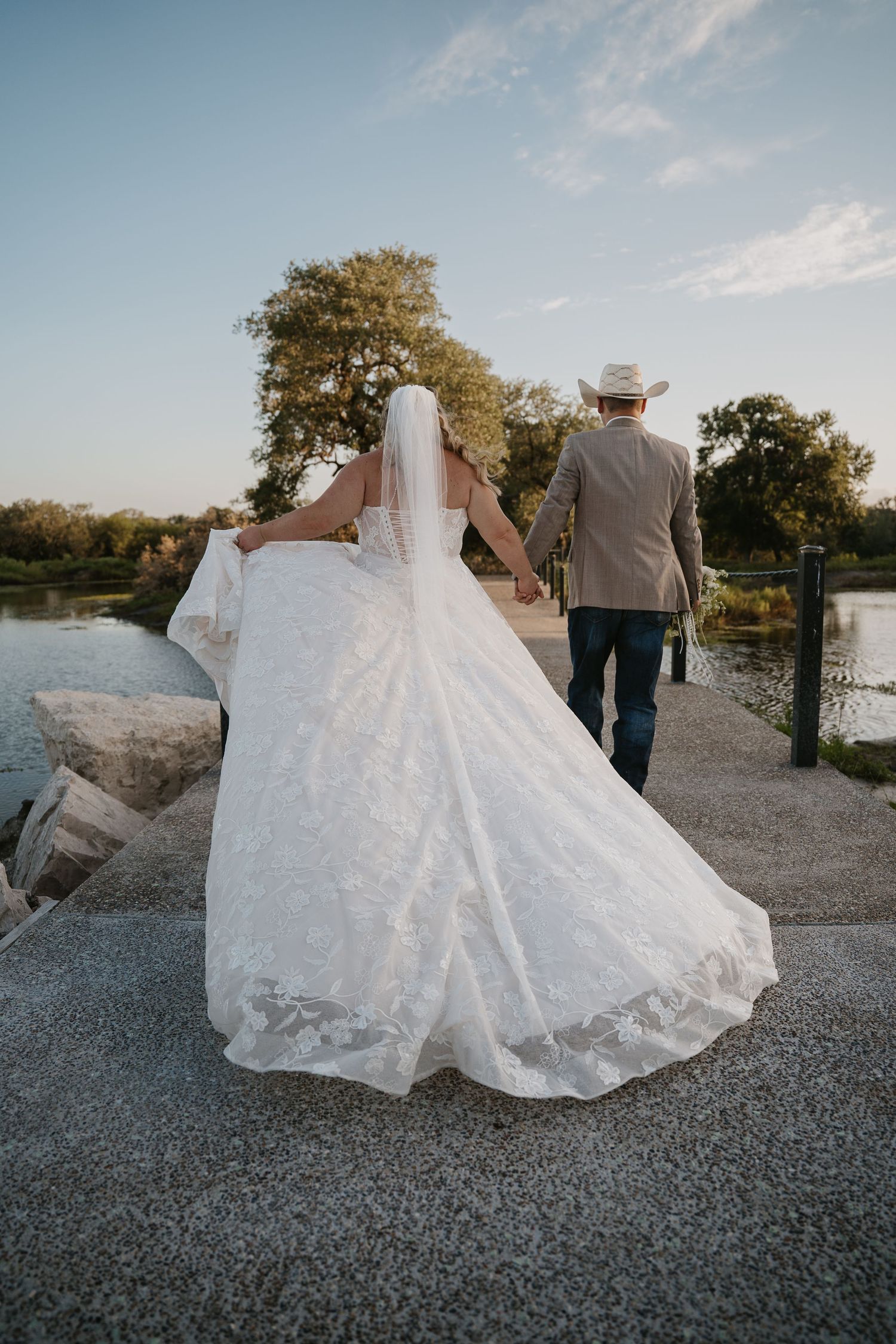Bride and groom in cowboy hat walk hand in hand along waterside path at sunset.