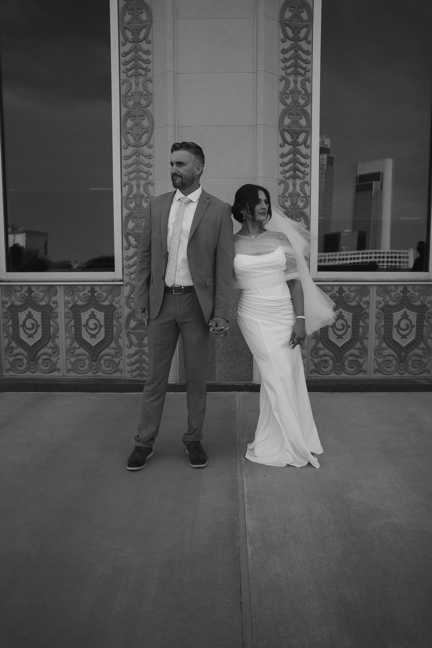 Elegant bride and groom hold hands in black and white wedding portrait against ornate building.