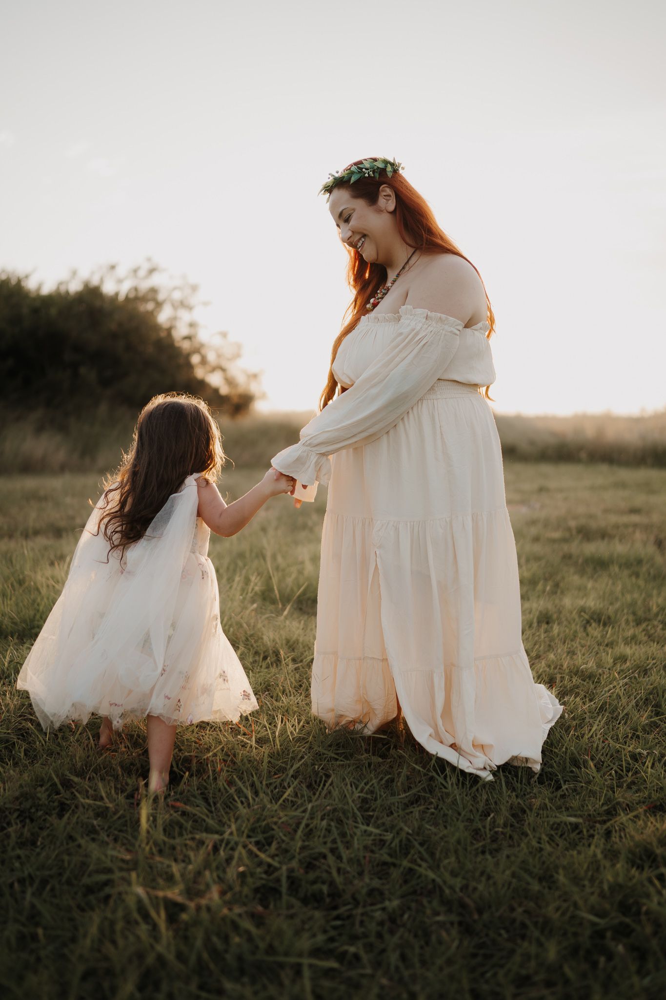 Woman in white boho dress holds daughters hand in golden sunset field.