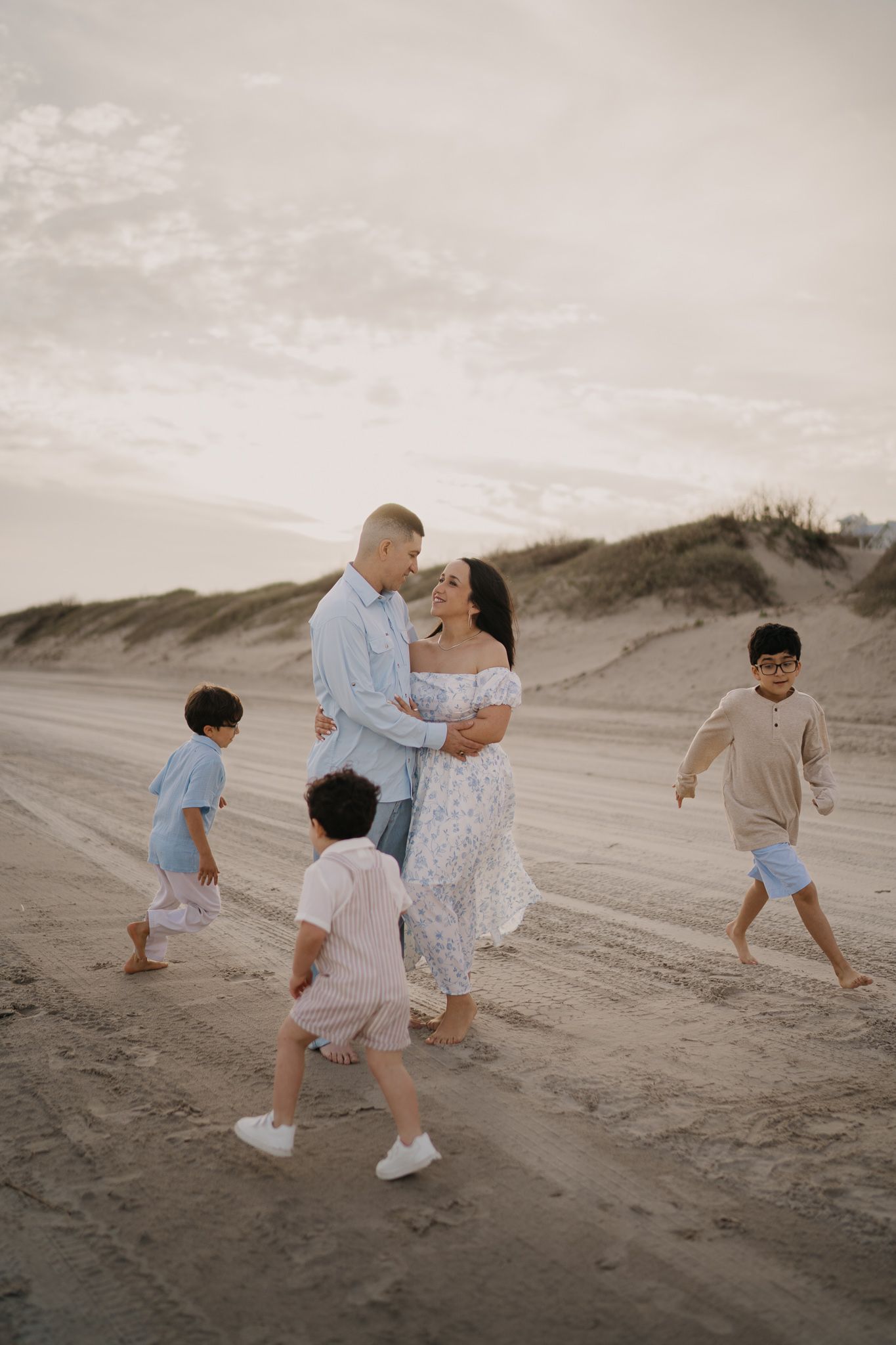 Family with three young boys walking barefoot on a sandy beach at golden hour, parents embracing in soft neutral tones.