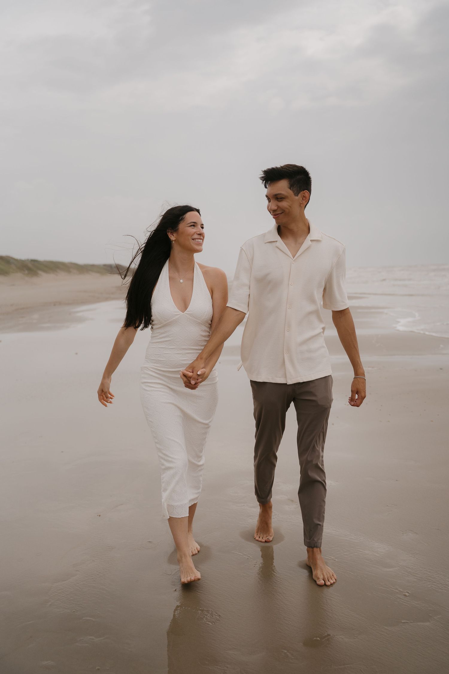 Couple holding hands walking barefoot on a beach, woman in white dress, man in white shirt and gray pants.