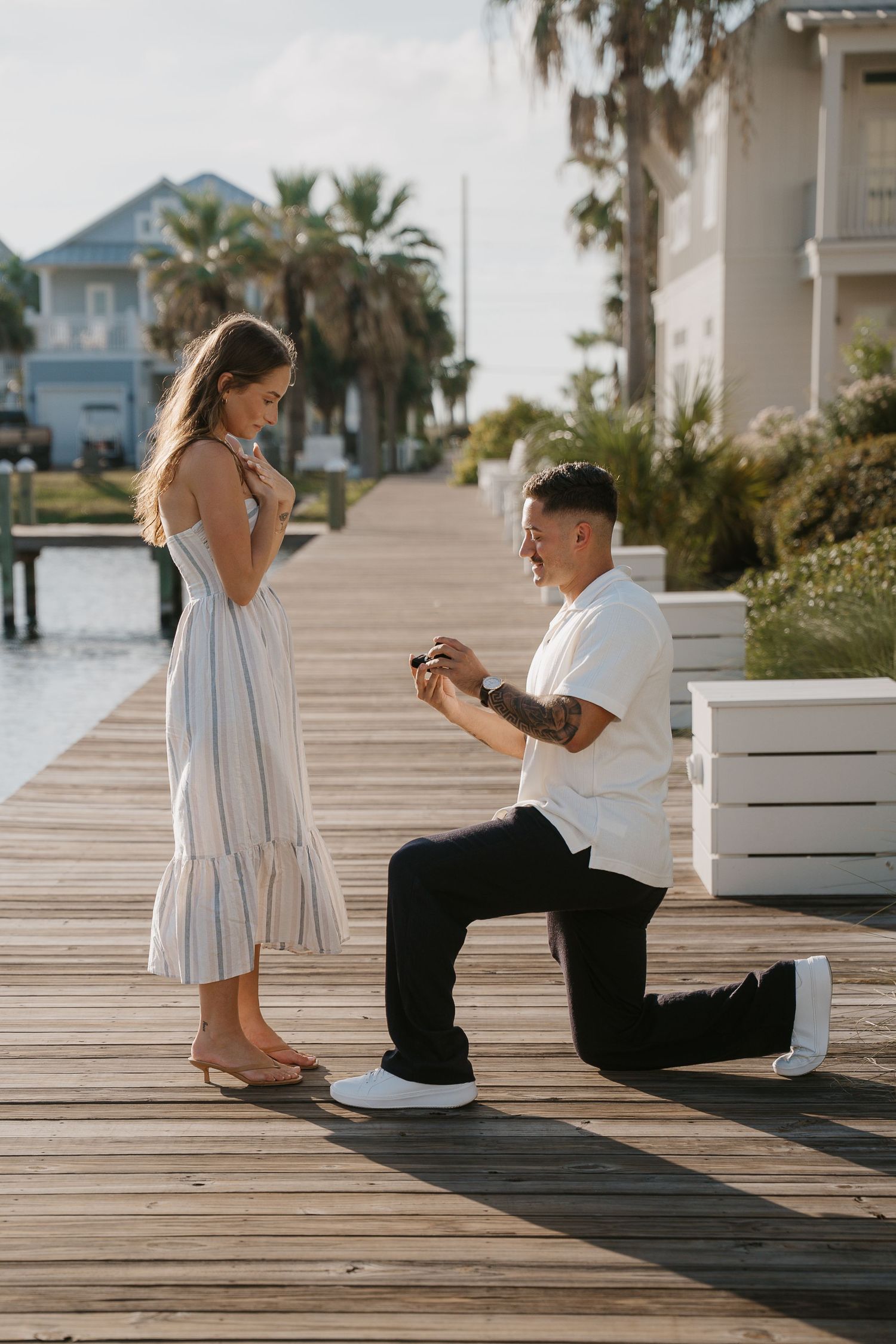 Man proposes on one knee to woman on a waterfront dock, holding a ring box, with palm trees and coastal homes in background.