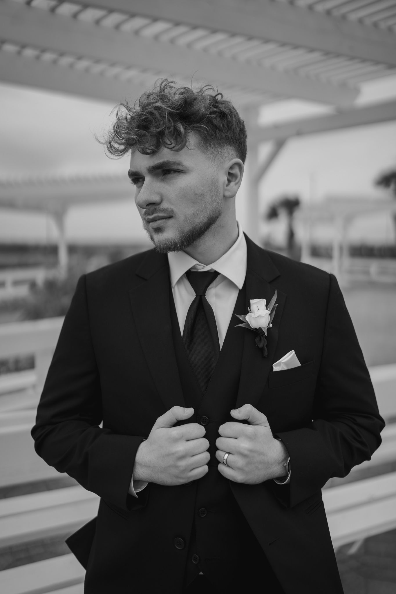 Black and white portrait of groom in black suit with boutonniere adjusting jacket outdoors.
