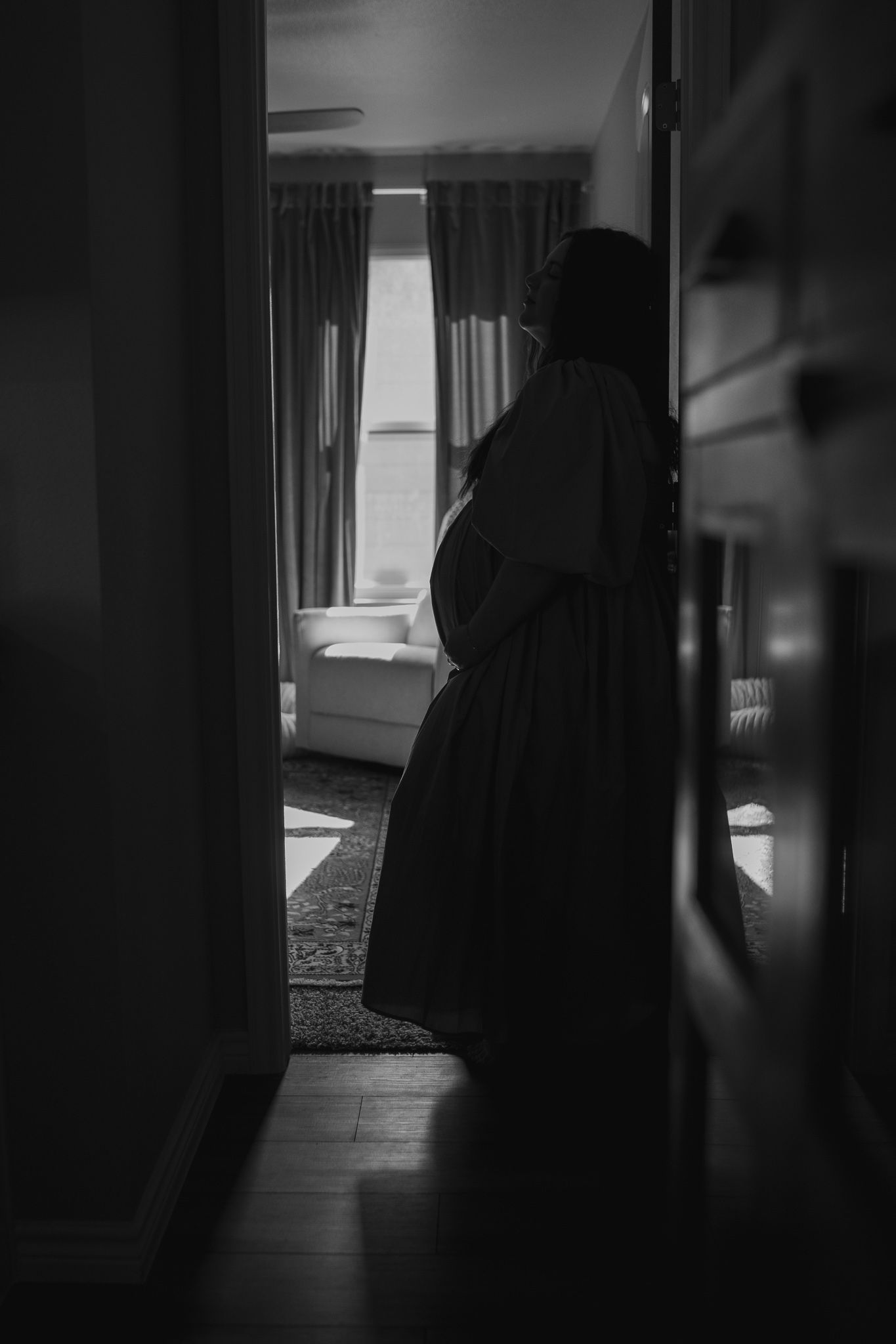 Silhouette of pregnant woman standing in doorway with natural window light in black and white.