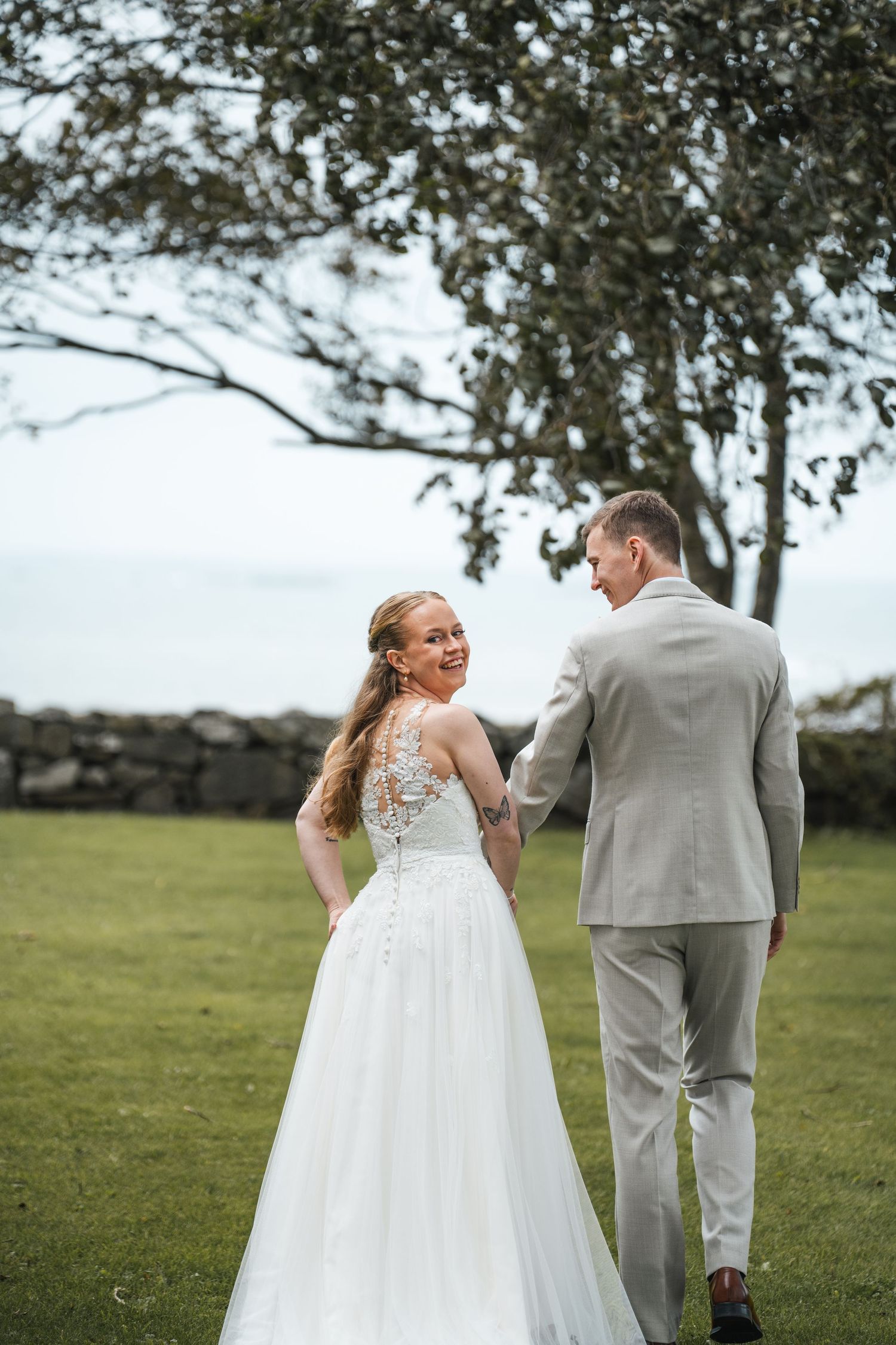 Bride looks back smiling at groom while walking on green lawn near stone wall.