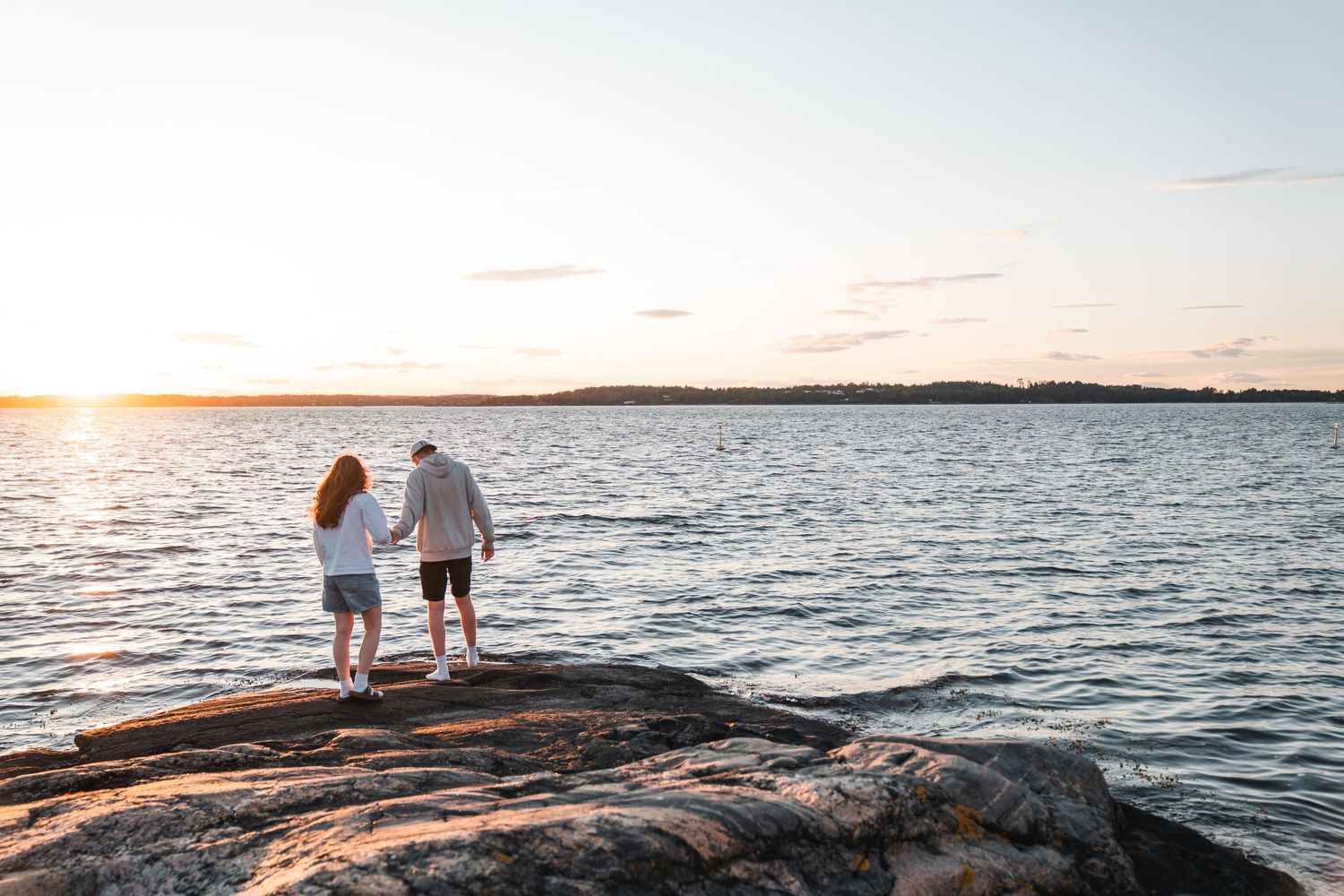 Couple holding hands standing on rocky shore watching a golden sunset over calm water.