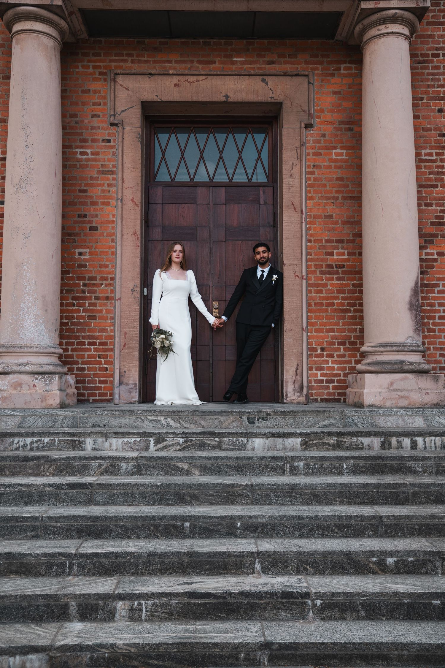 Bride and groom holding hands on steps of a brick church with tall columns.