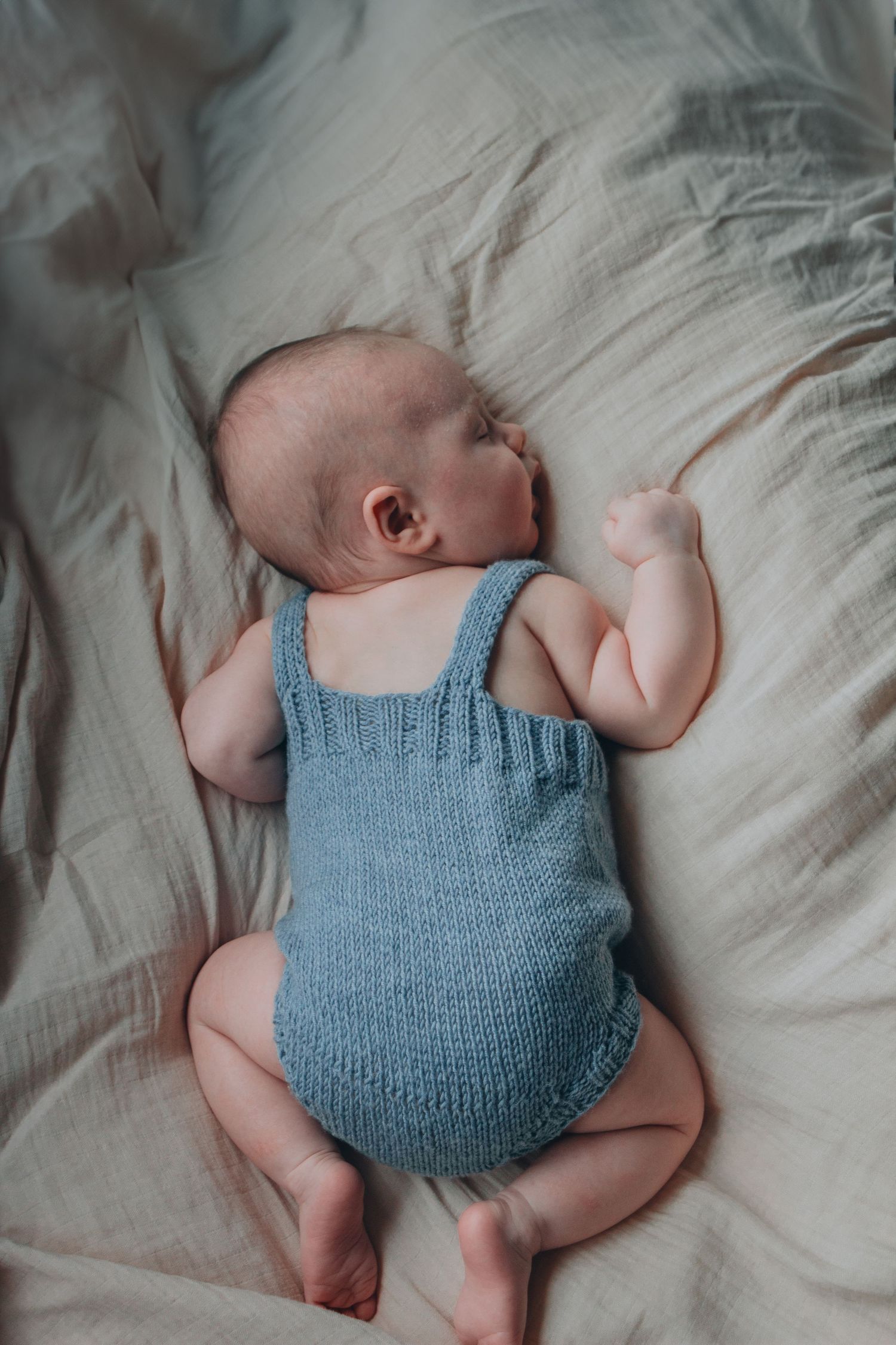 Newborn baby in a blue knitted romper lying on their tummy on a beige linen blanket.