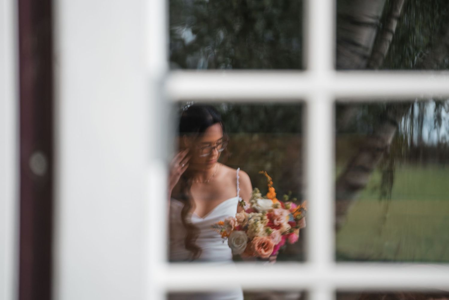 Bride in white dress holding colorful bouquet photographed through white window panes.