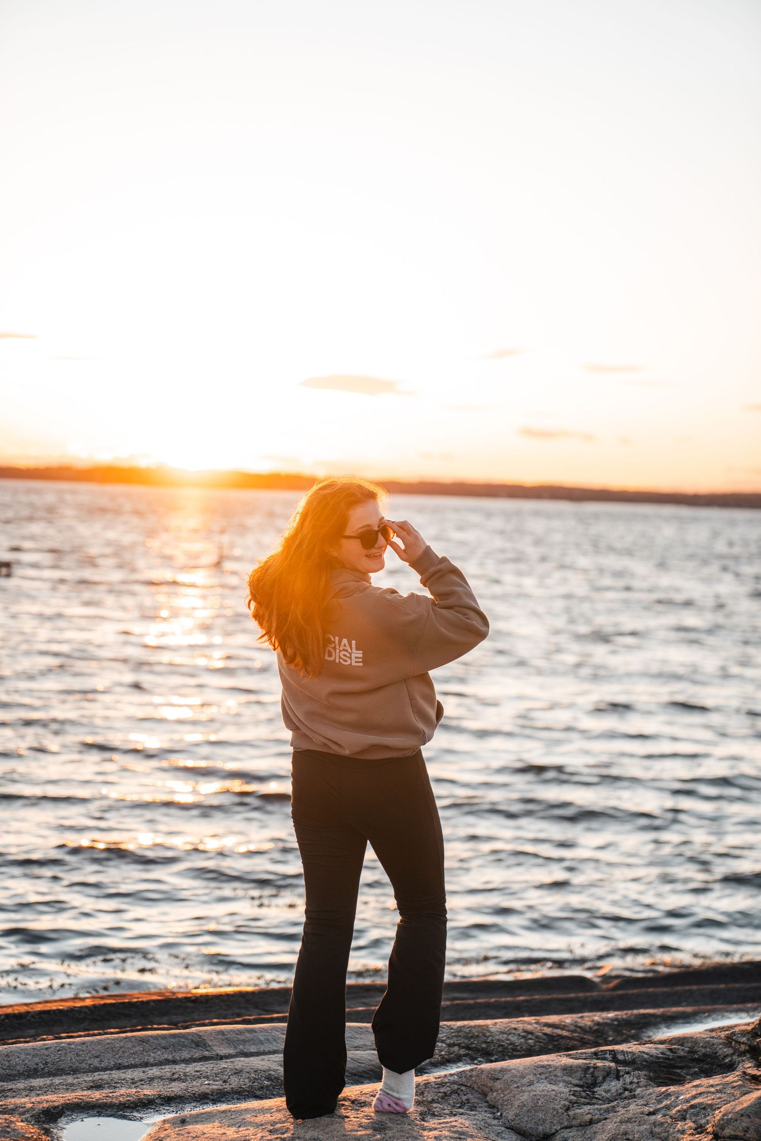 Young woman in gray hoodie and black flares posing by water at golden sunset.