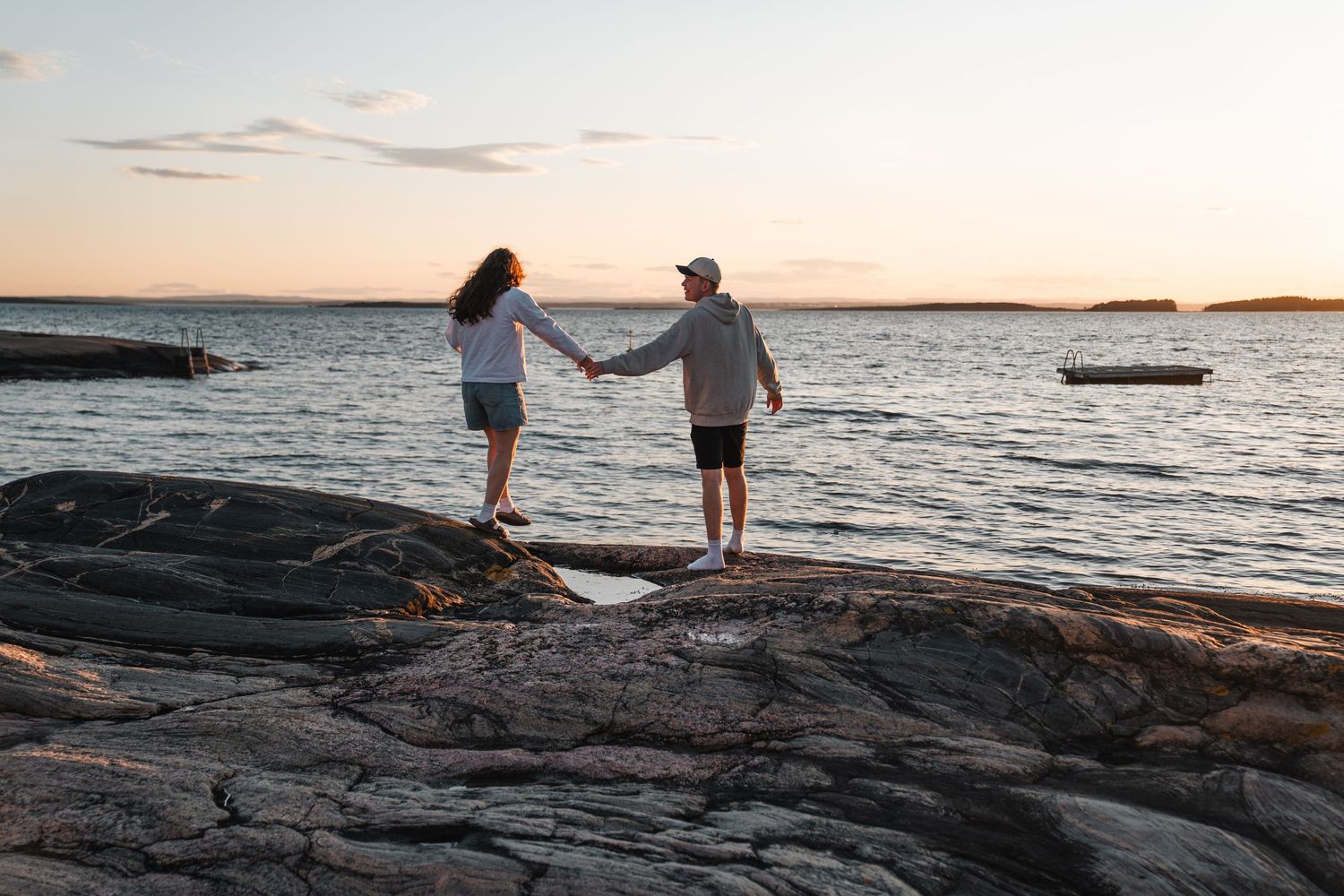 Couple holding hands on rocky shoreline at sunset with calm ocean view.