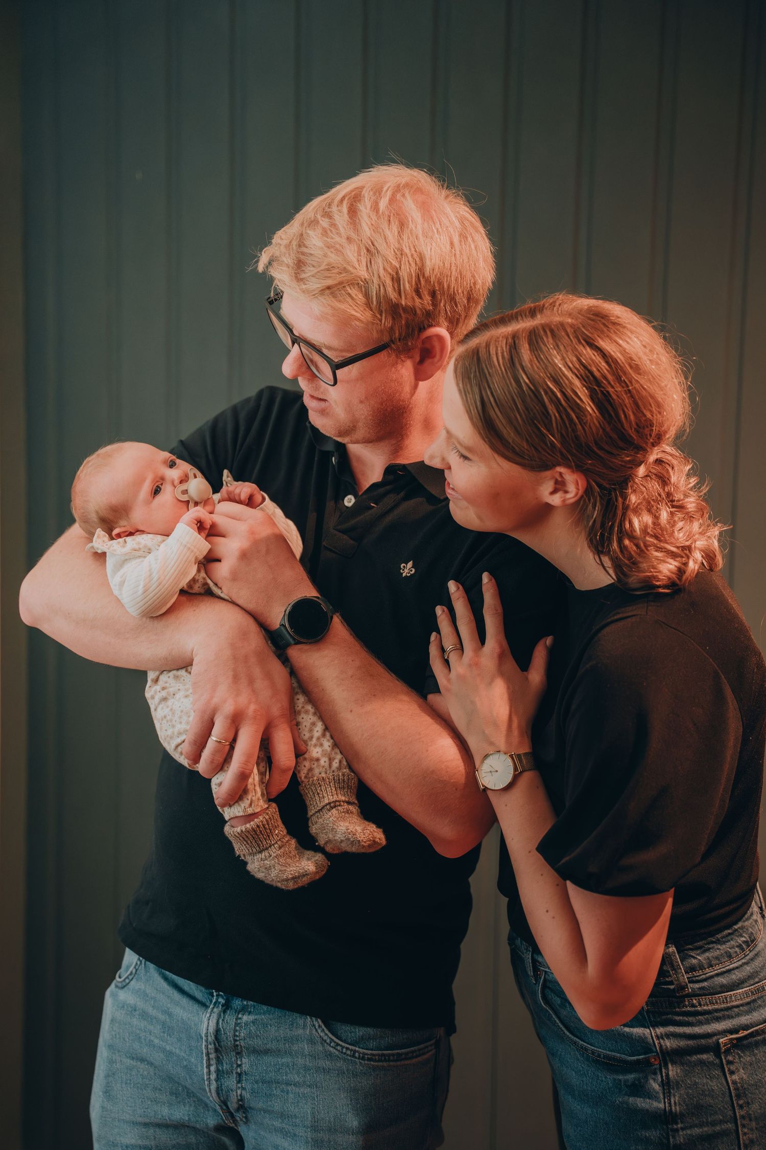 Red-haired couple in black shirts lovingly admiring newborn baby in indoor portrait session.
