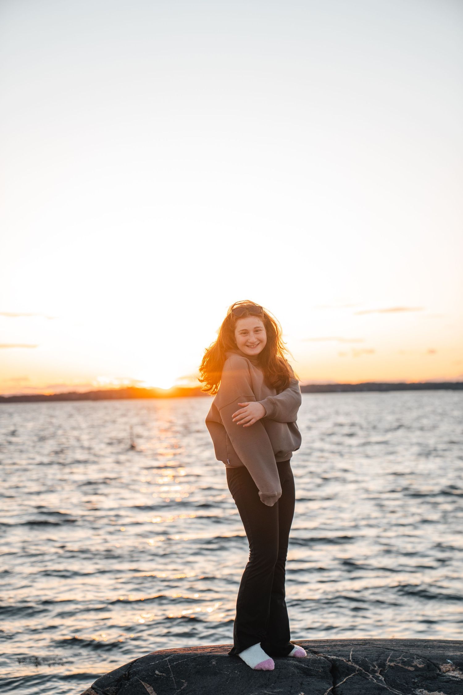 Young woman with curly hair standing on rocks smiling at sunset by the water.