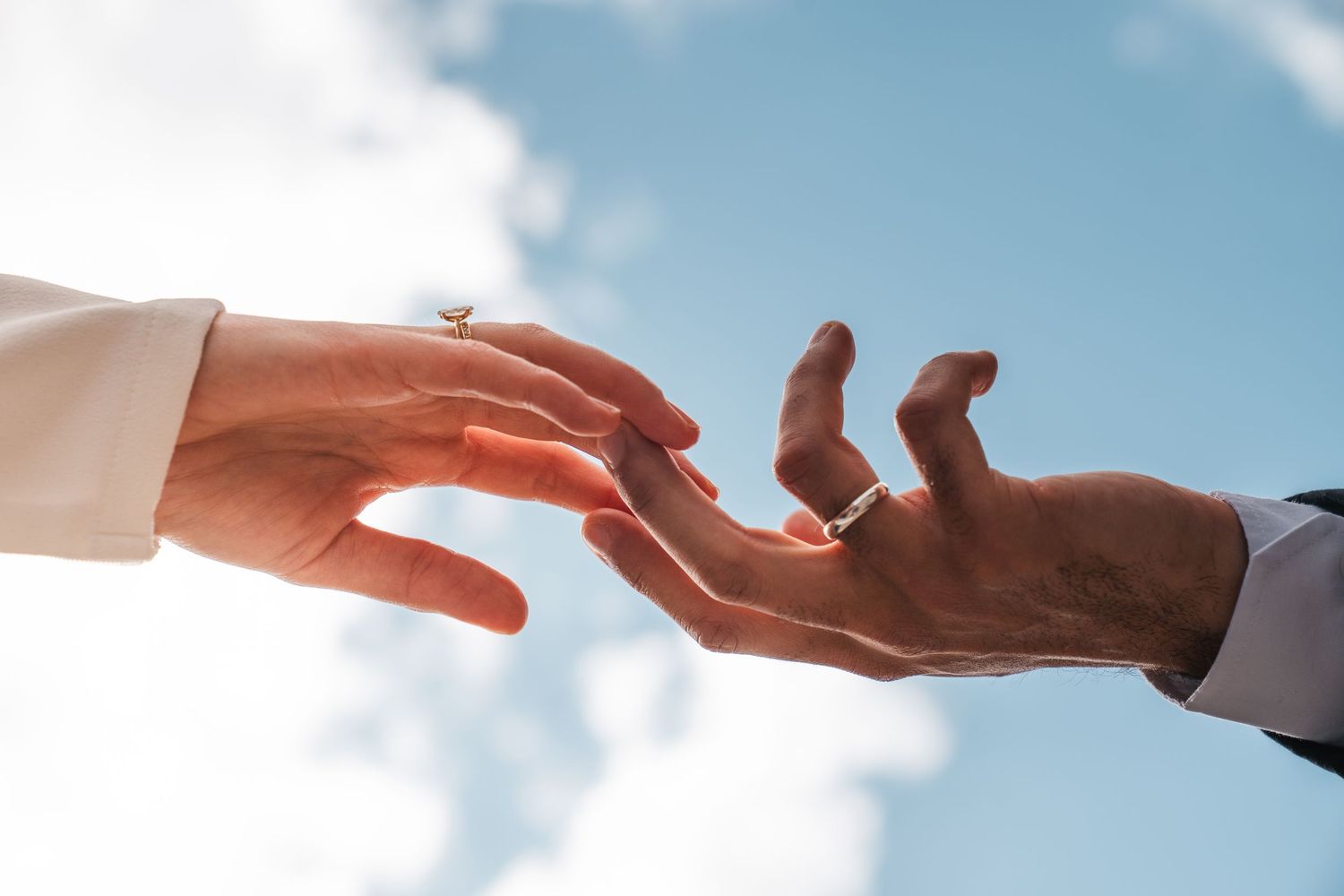 Two hands reaching toward each other with wedding rings against a blue sky.