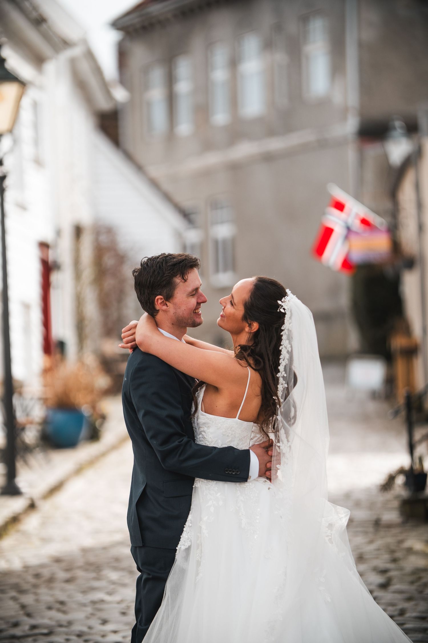 Bride and groom embrace on cobblestone street with Danish flag in background.