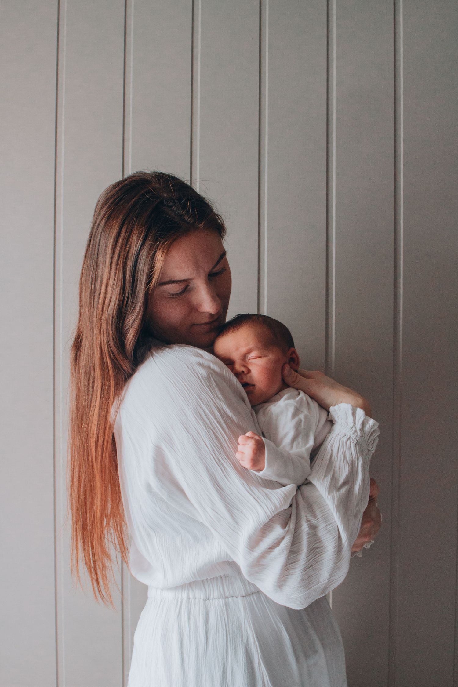 A mother in a white dress tenderly cradling her sleeping newborn baby against a grey wall.