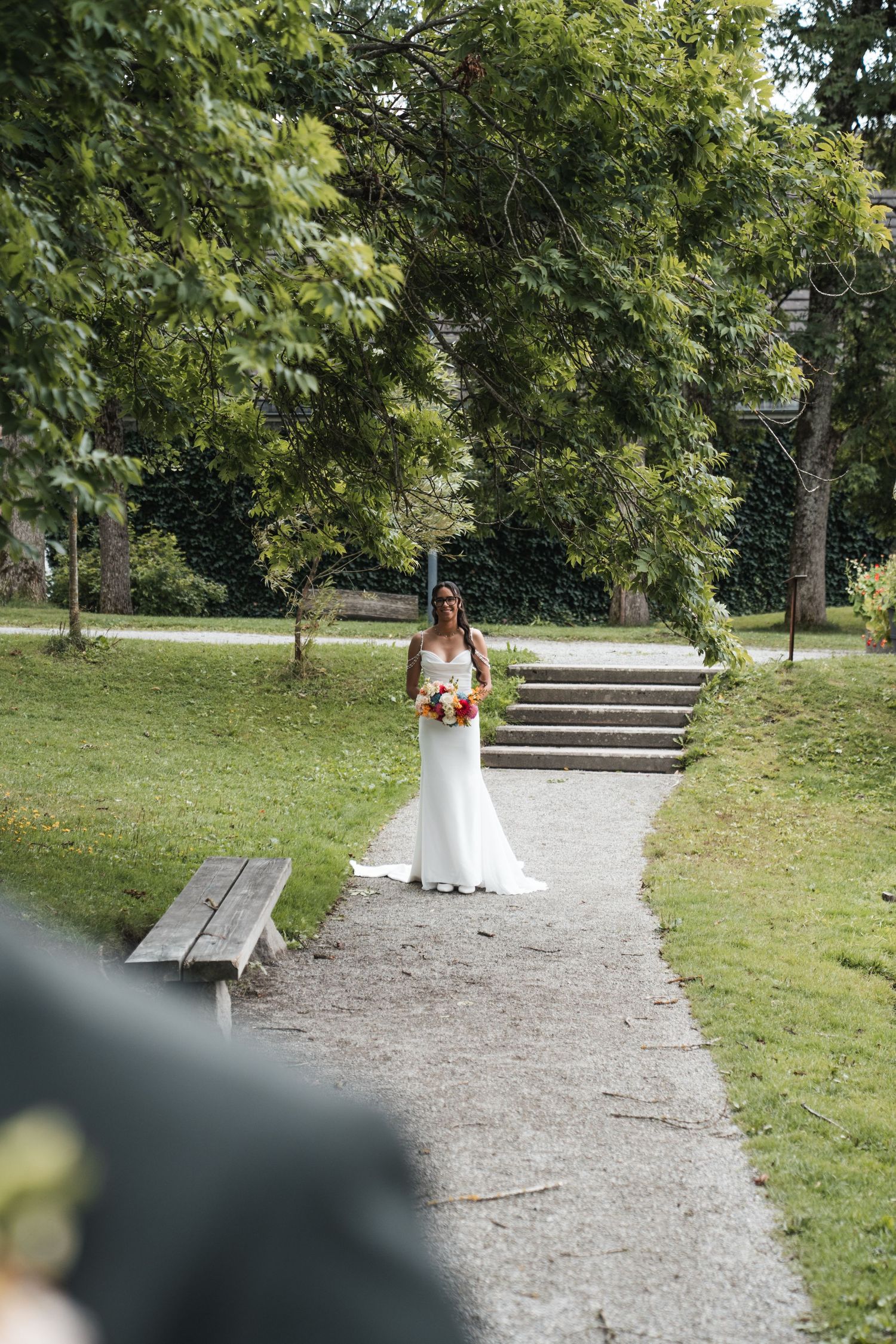 Bride in white gown holding colorful bouquet standing on garden path surrounded by lush green trees.