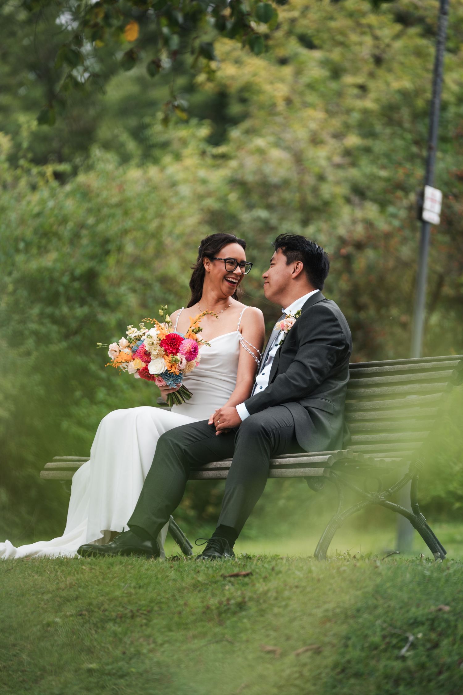 Bride and groom sitting on a park bench laughing together with a colorful bridal bouquet.