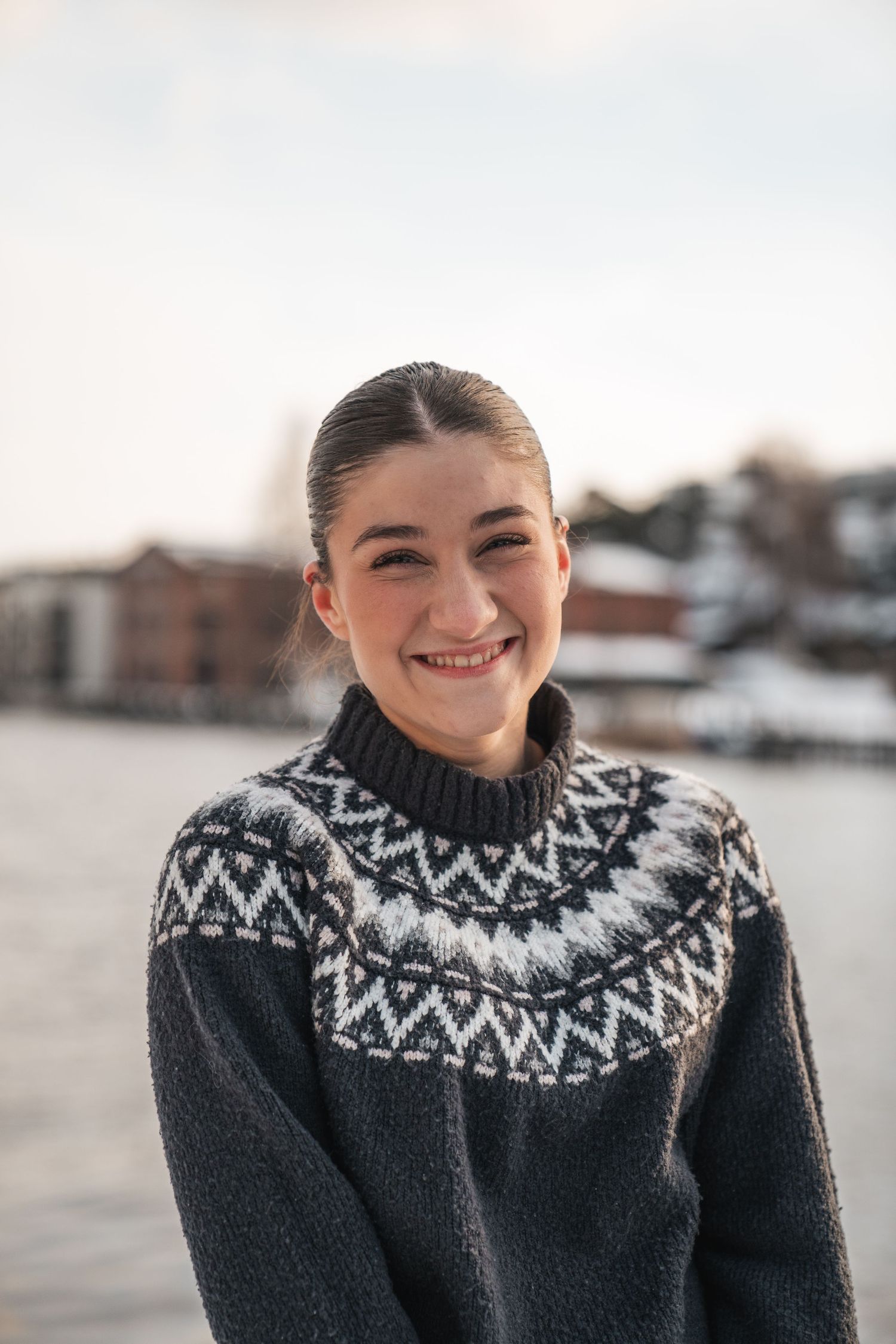 Smiling young woman in Nordic patterned sweater posing outdoors by waterfront in winter.