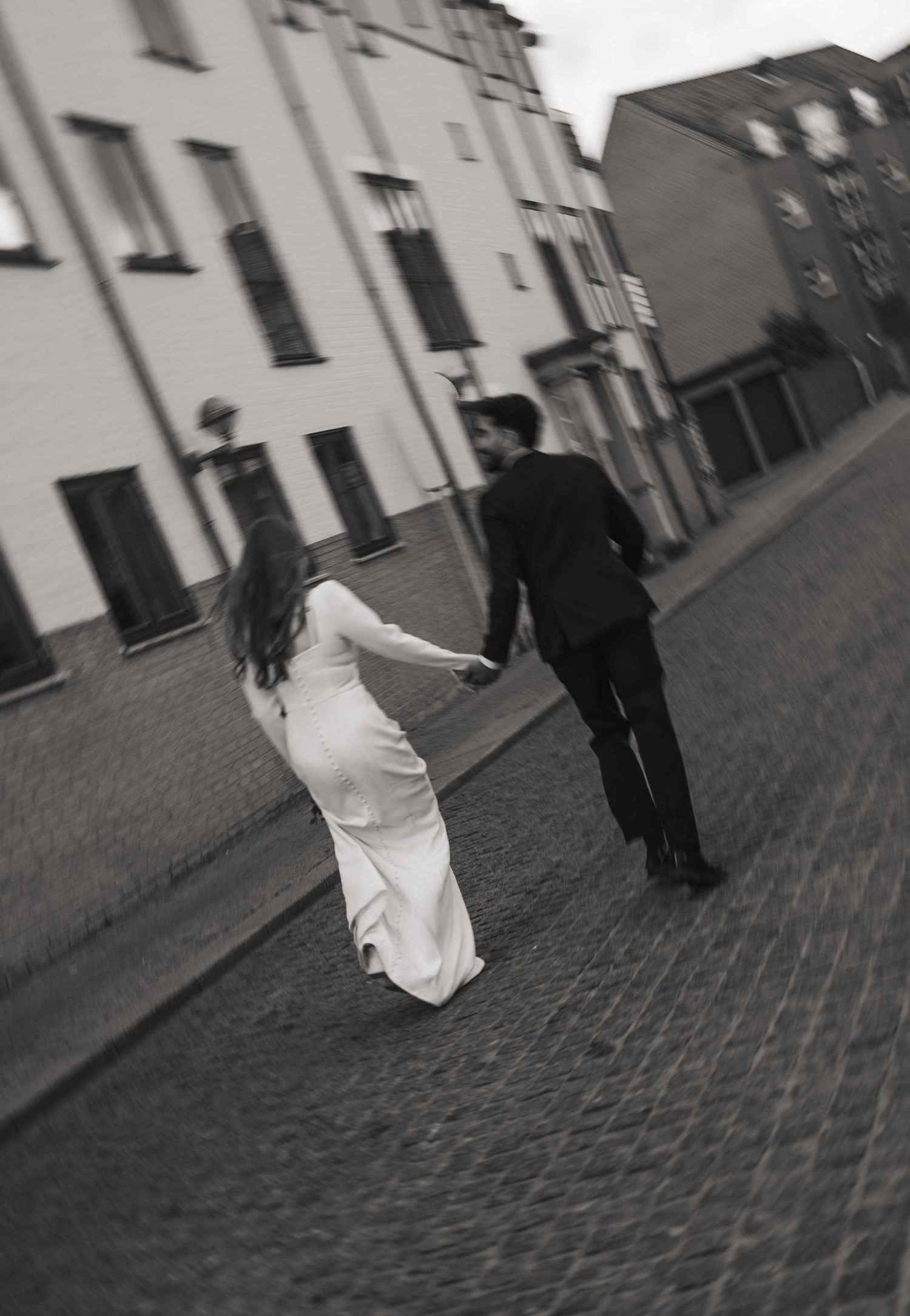 Black and white photo of bride and groom holding hands walking on cobblestone street.