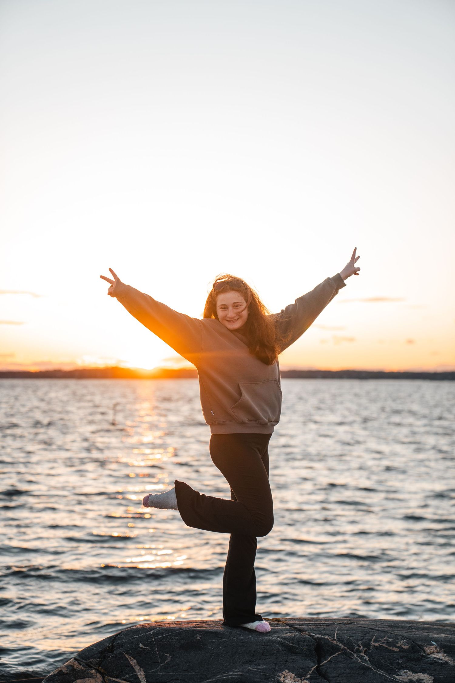 Young woman balancing on one leg with arms raised joyfully at a lakeside sunset.