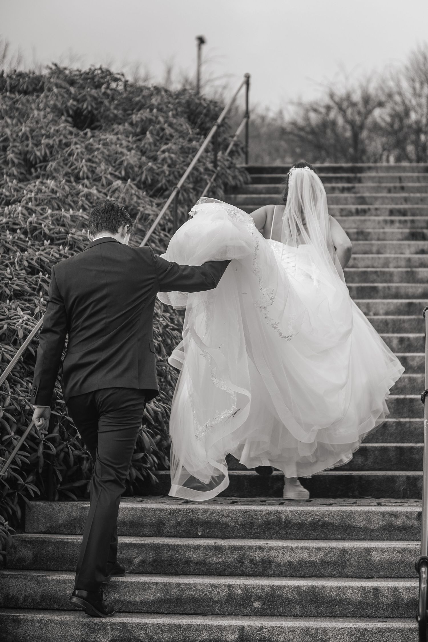 Black and white photo of groom helping bride carry her dress up outdoor stone steps.