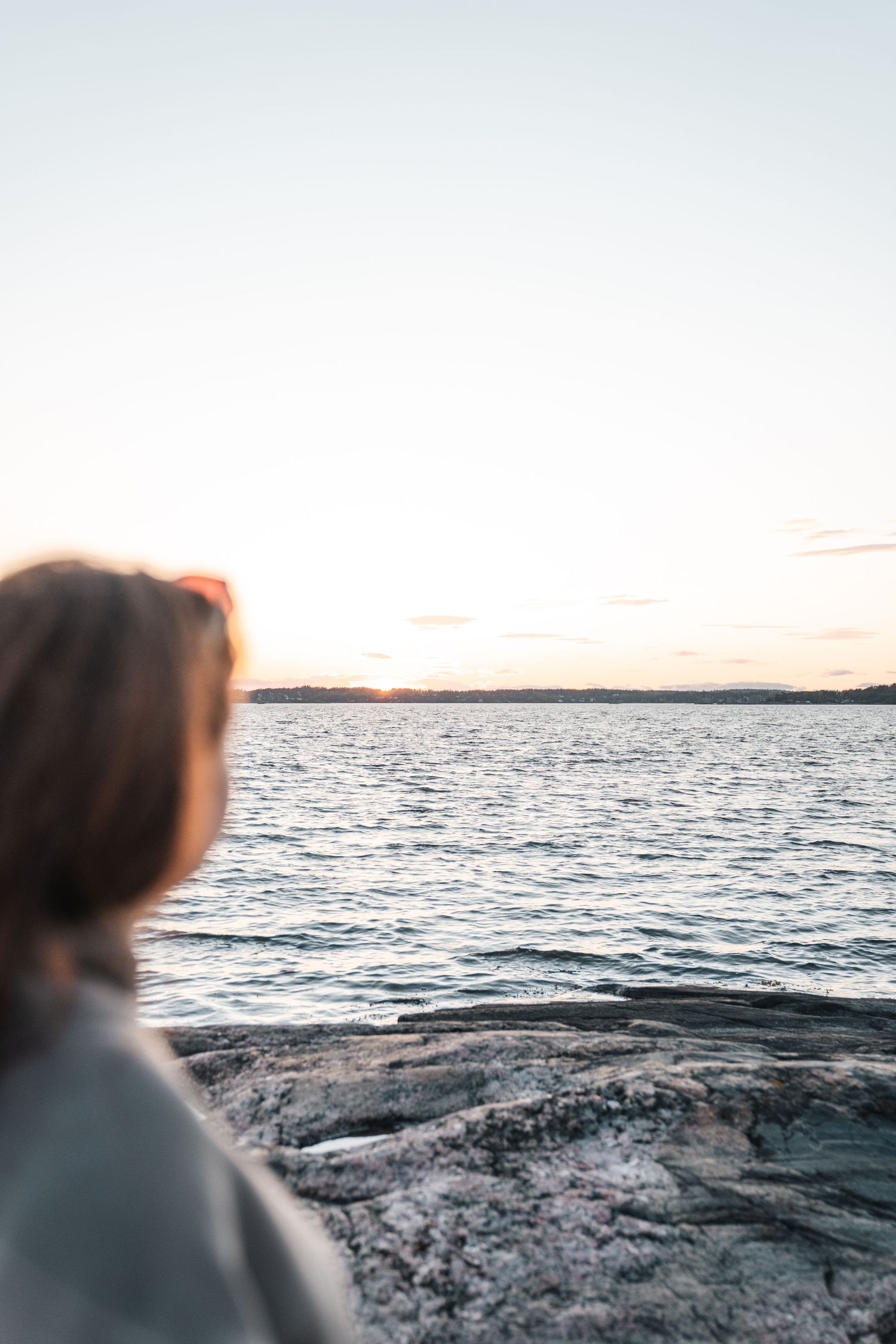 Woman sitting on rocky shore watching a serene sunset over calm lake waters in golden hour light.