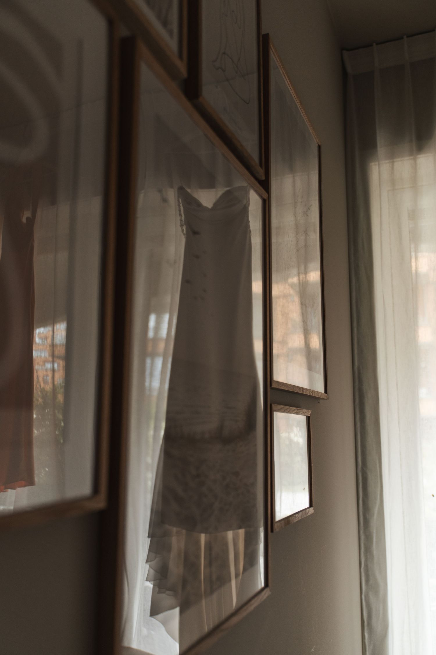 Elegant strapless wedding dress hanging near window with soft natural light and framed pictures on wall.