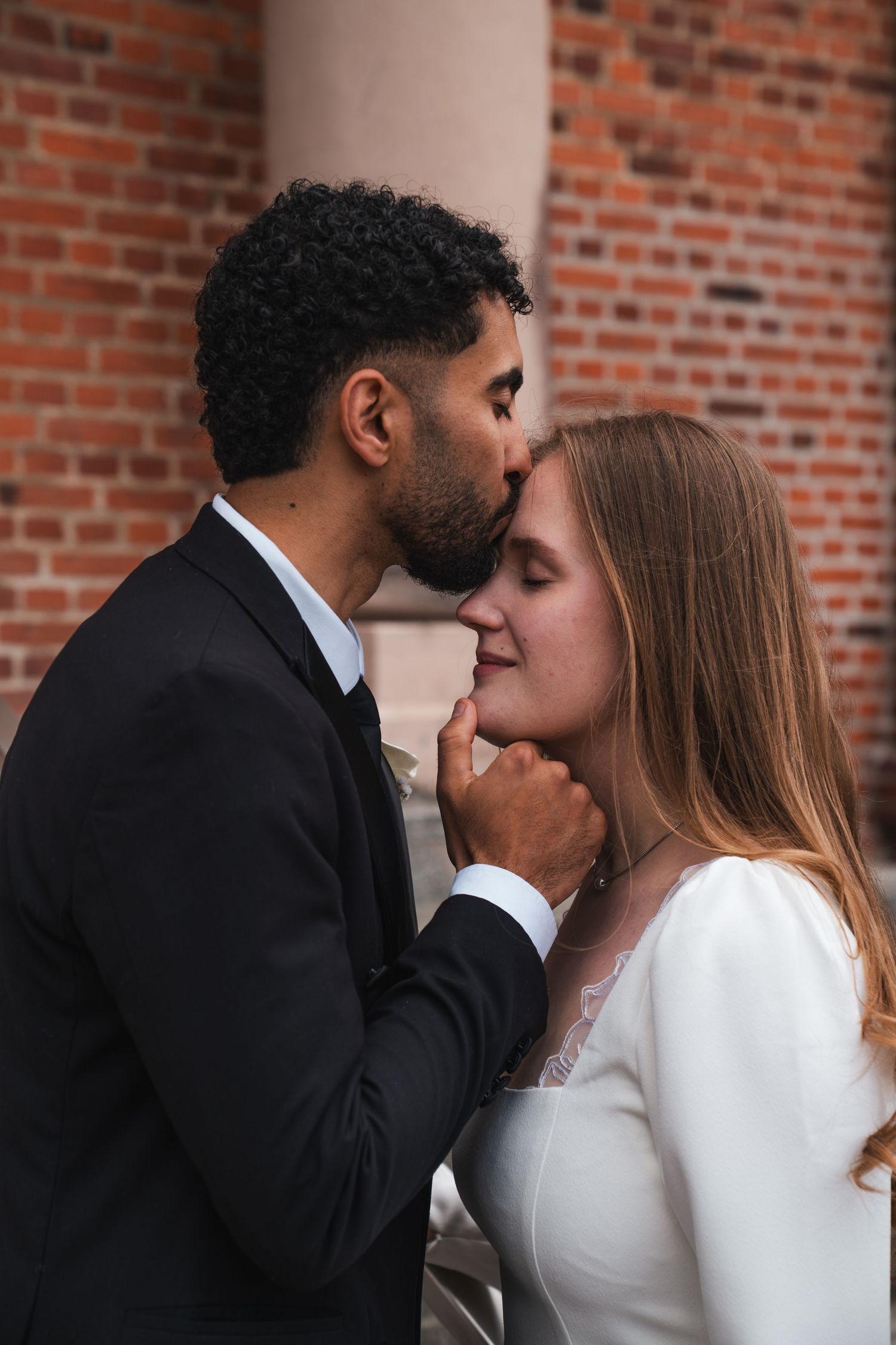 Groom in black suit kissing bride on forehead against brick wall backdrop.