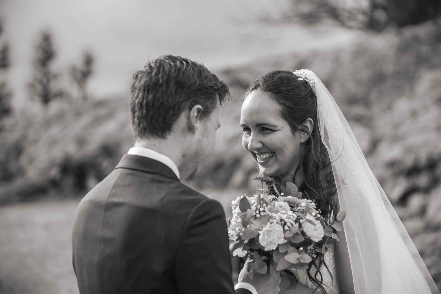 Black and white wedding photo of smiling bride holding bouquet facing groom outdoors.