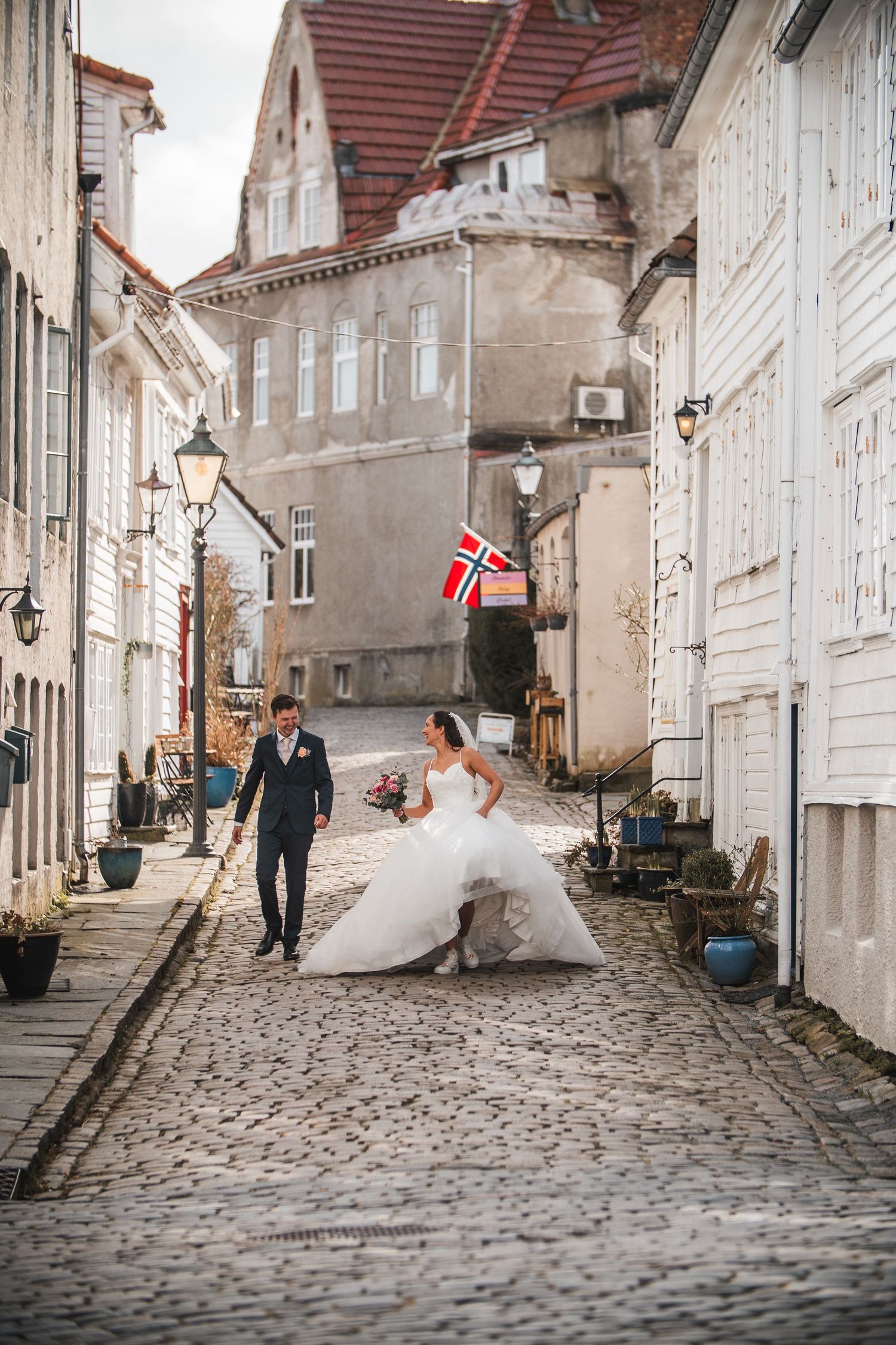 Wedding couple walks cobblestone street in Norway with Norwegian flag visible.