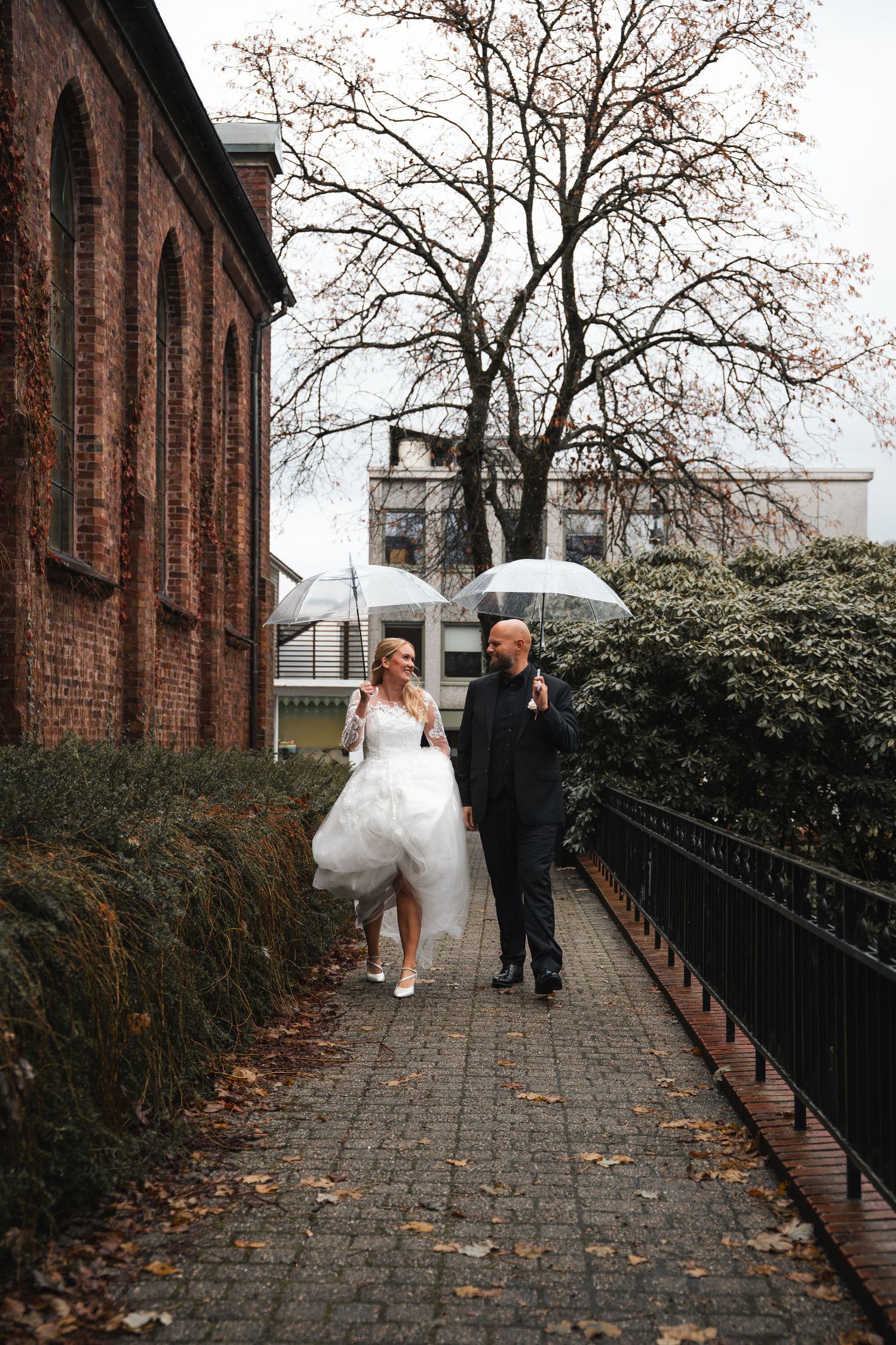 Bride and groom walk with clear umbrellas on rainy autumn day near brick church.