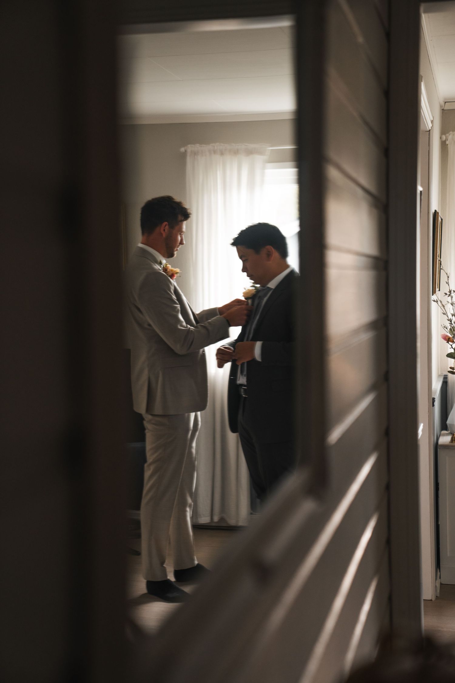 Groom adjusts groomsman tie in sunlit room, viewed through doorway.
