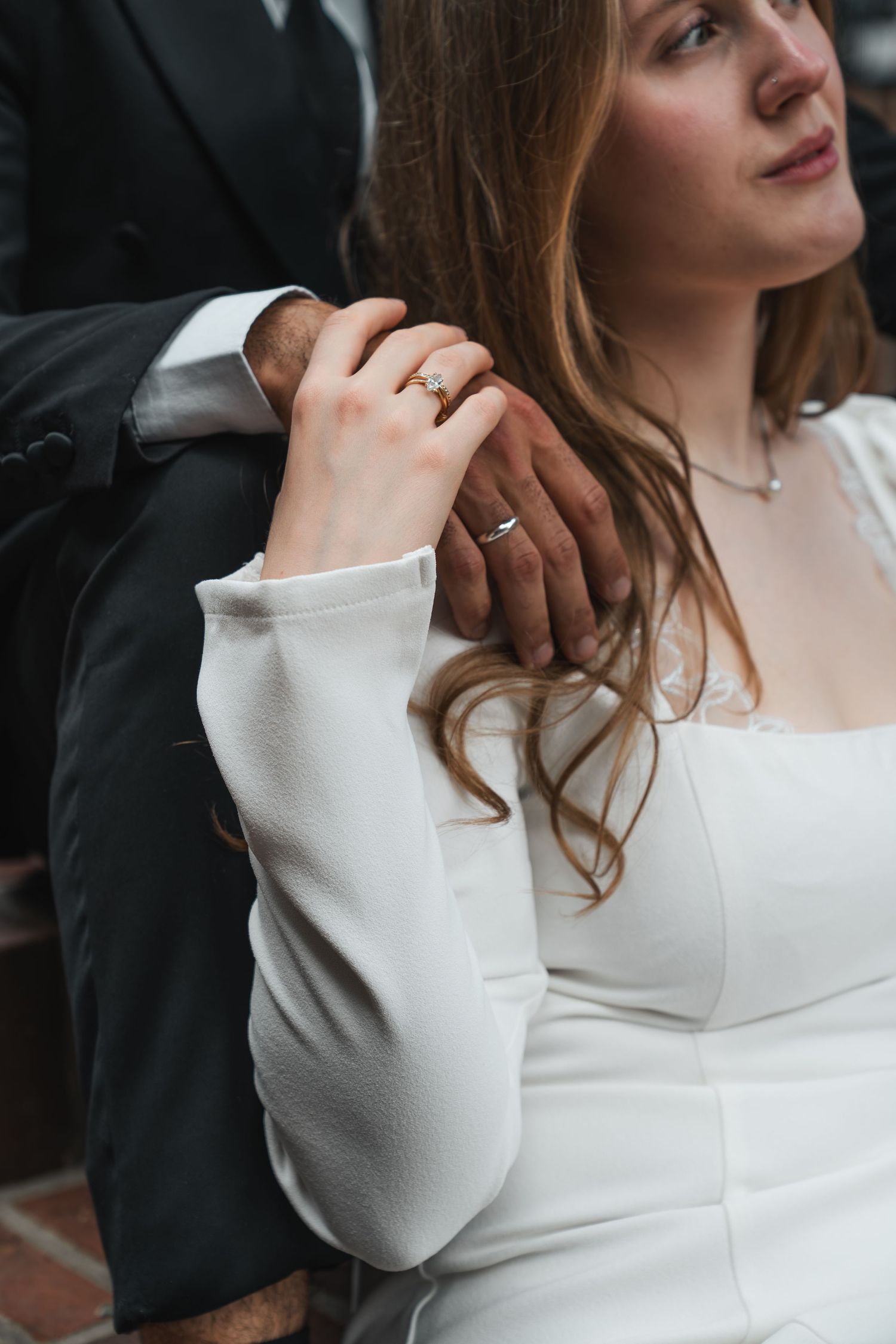 Close-up of bride in white dress holding grooms hand with engagement ring visible.