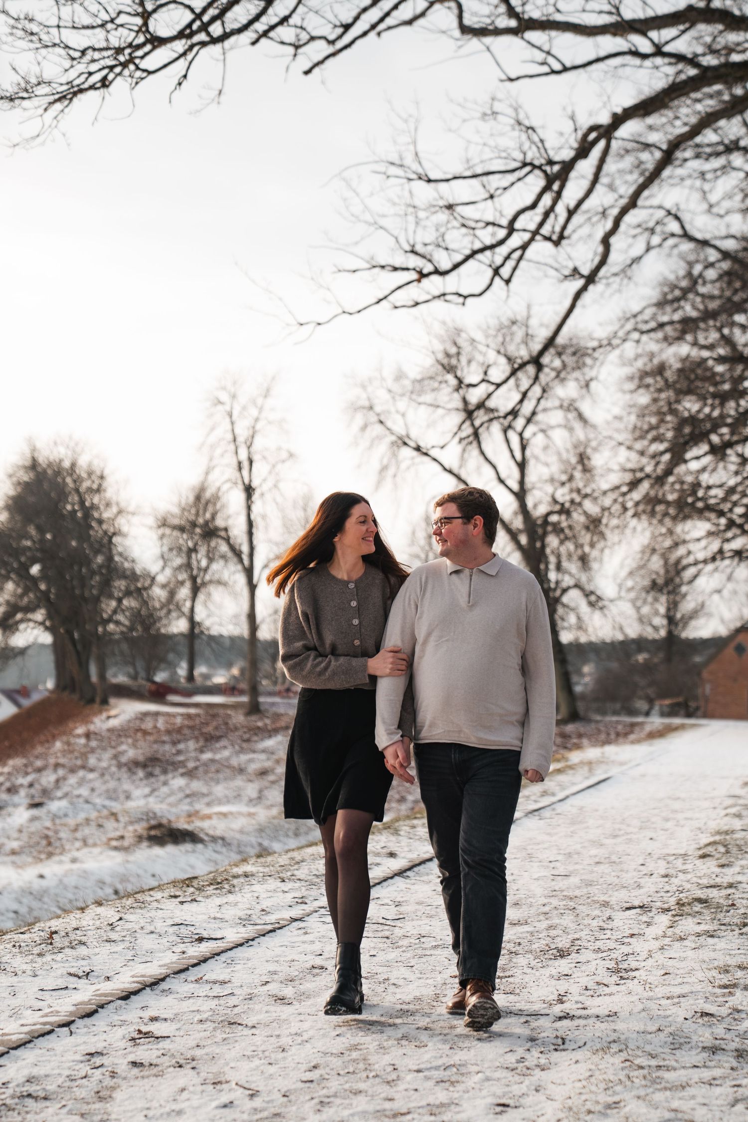 Happy couple walking hand in hand along a snowy path surrounded by bare winter trees.