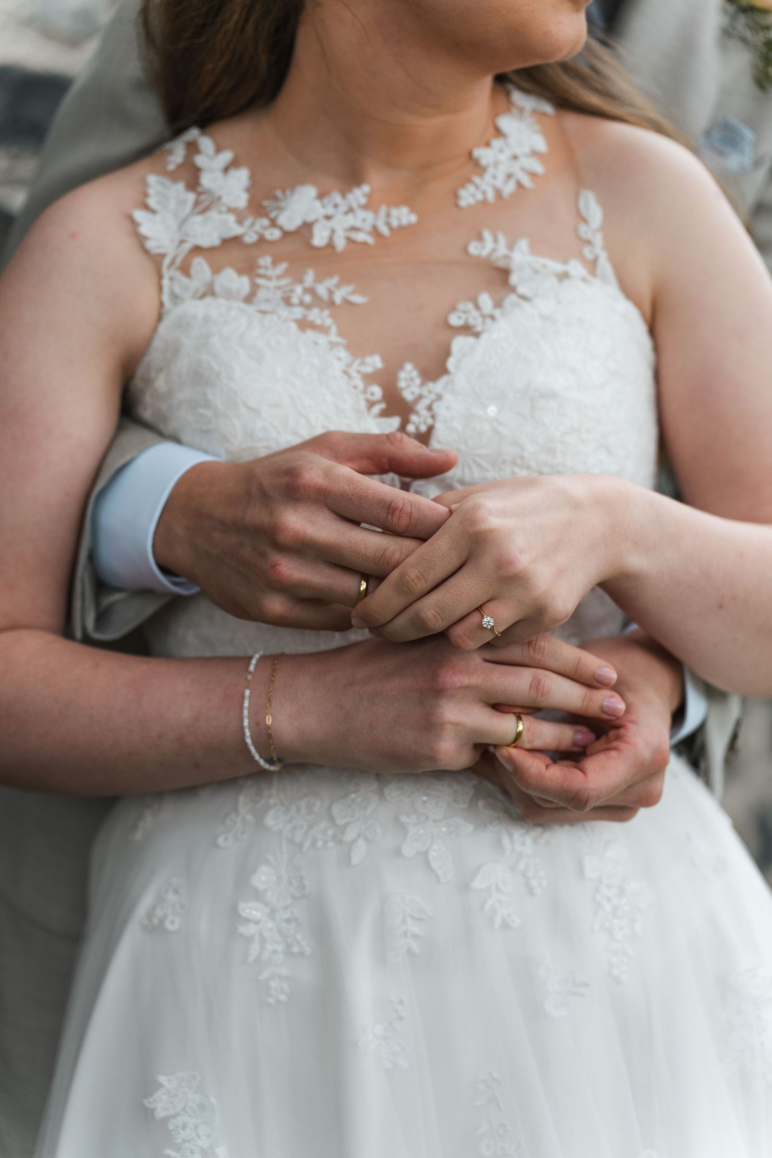 Close-up of bride and groom holding hands showing wedding rings and lace gown.