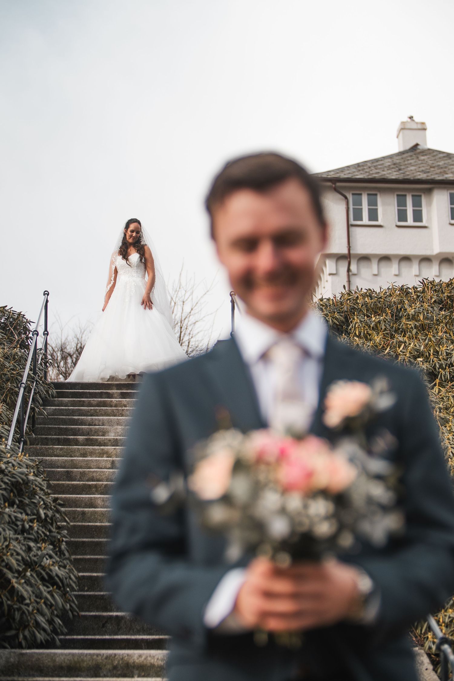 Smiling groom holding bouquet in foreground while bride descends outdoor staircase behind him.