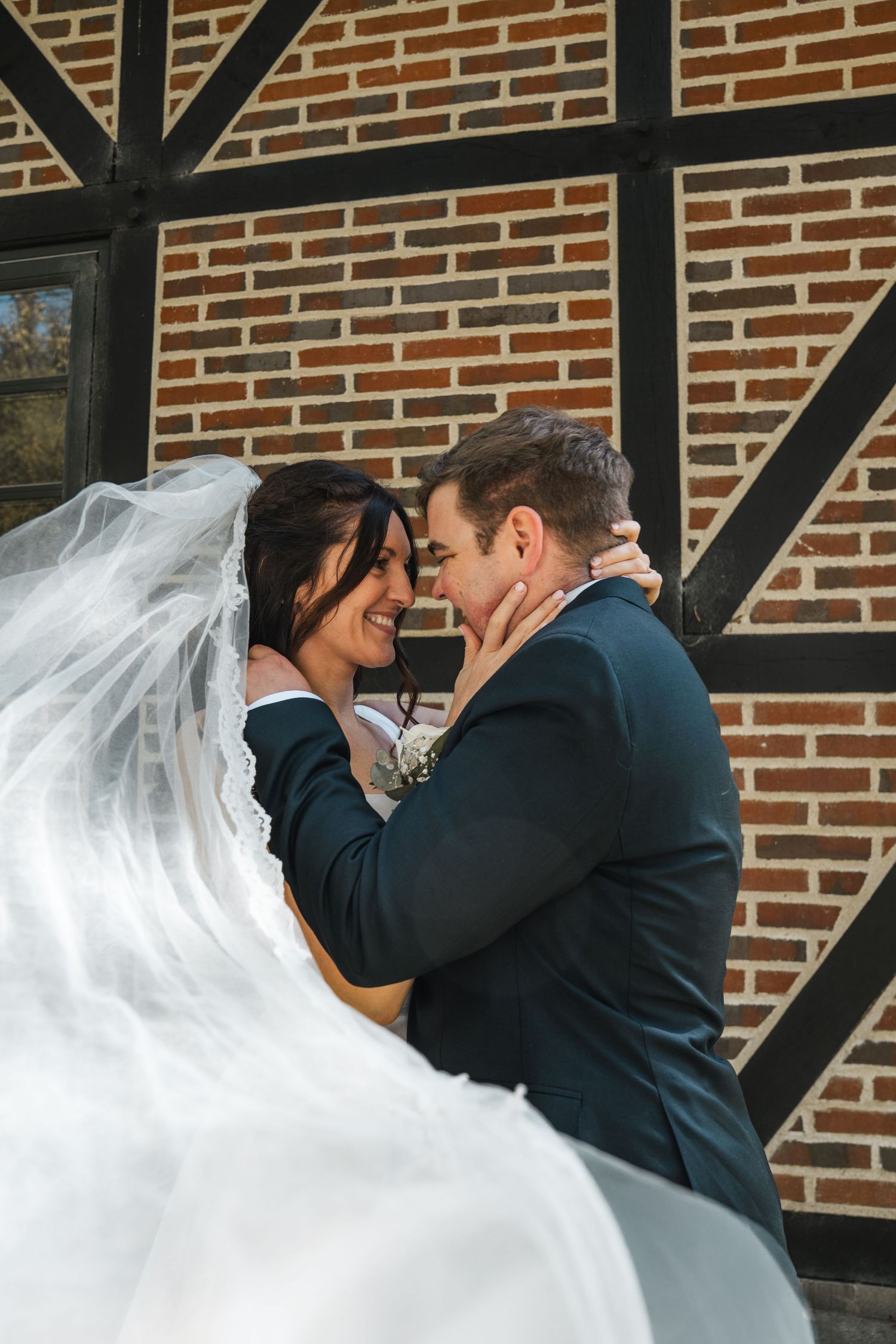 Bride and groom nearly kissing against brick wall, veil flowing in the wind.