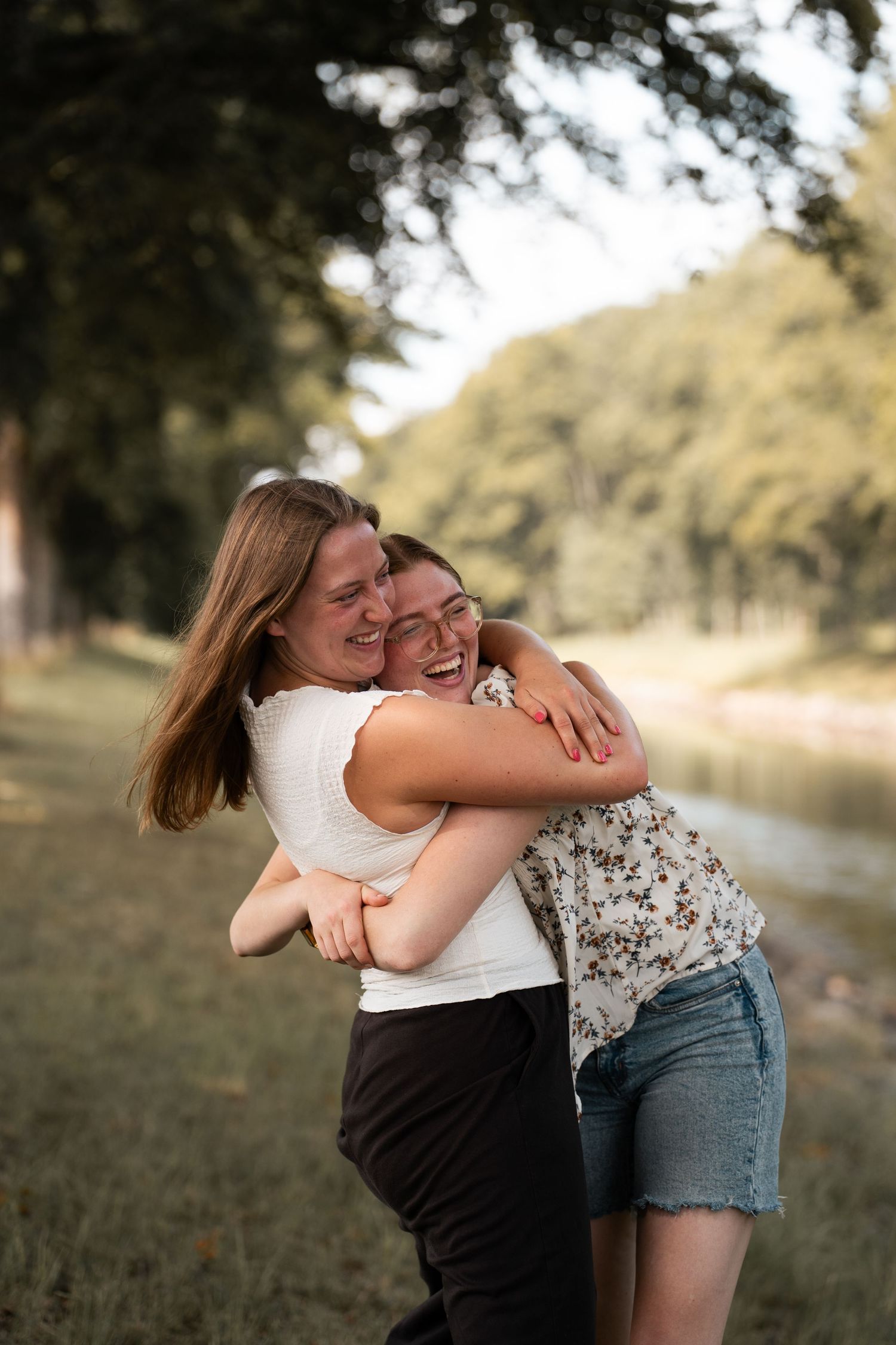 Two young women laughing and hugging each other in a sunny outdoor park setting.