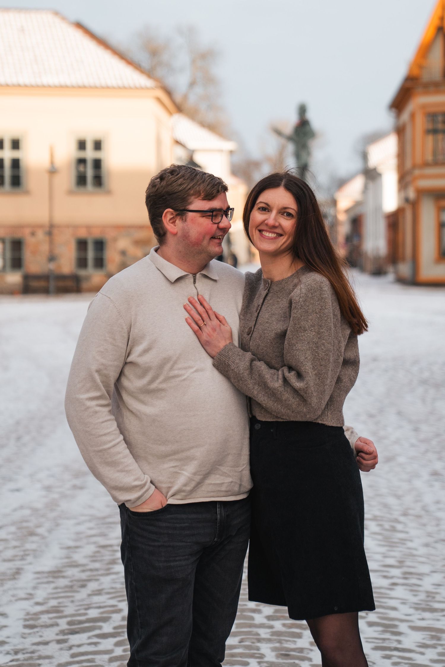 Smiling couple poses on snowy cobblestone square at golden hour in winter.