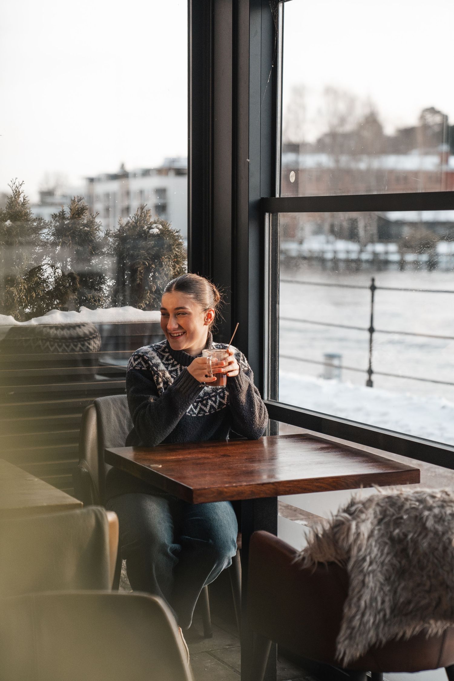 Happy woman in Nordic sweater sitting at cafe table by snowy window holding warm drink.